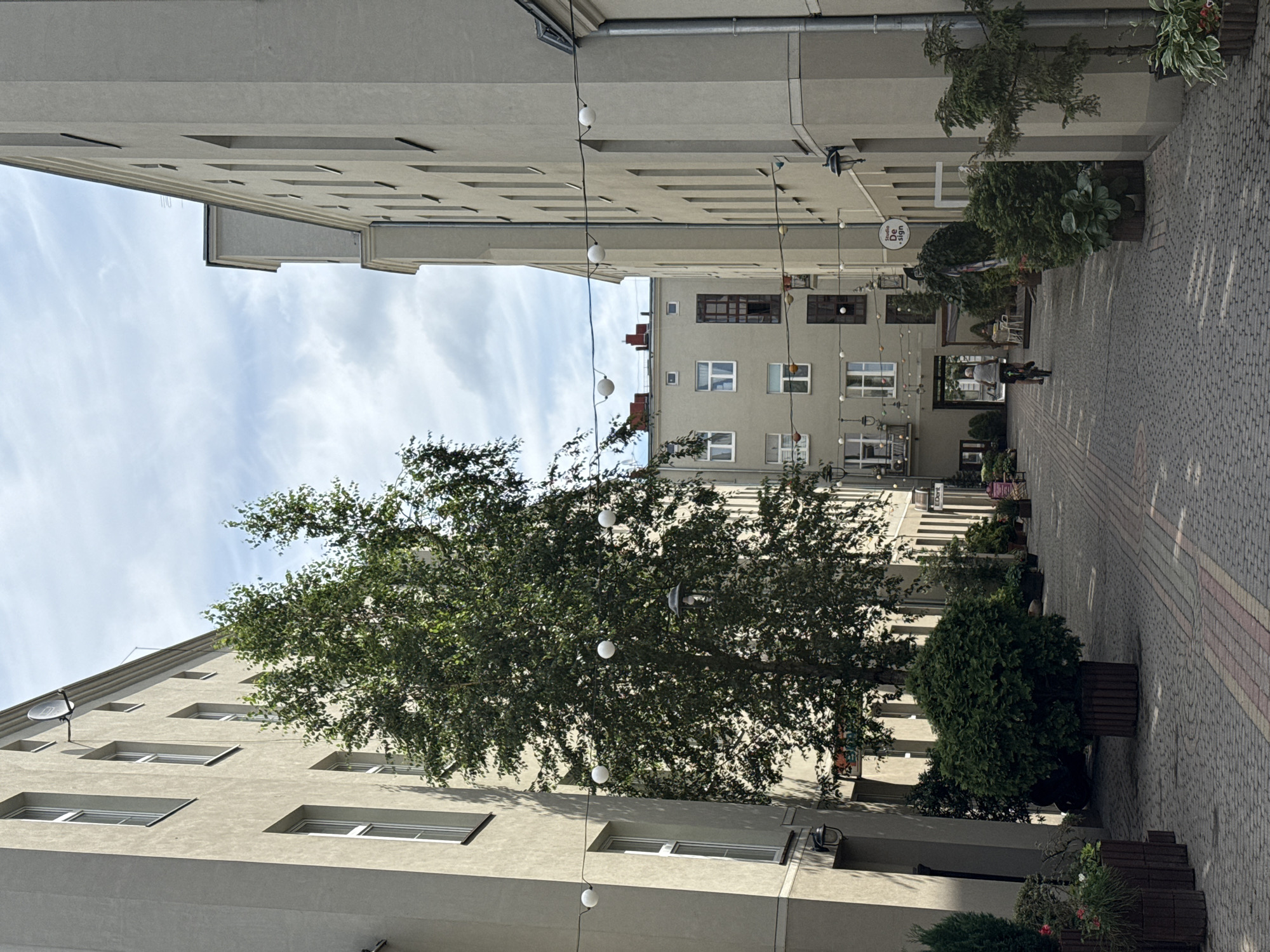 The image depicts a narrow street or courtyard flanked by multi-story buildings in Łódź, Poland. The architecture appears to be a mix of older, possibly industrial-era buildings, with some newer elements. The buildings are predominantly rendered in a light beige or grey color and have numerous windows. String lights with spherical bulbs are strung across the street, adding a decorative touch.
In the foreground, a person is riding a bicycle away from the camera. The street is paved with interlocking stones, with a pattern of alternating colors. Potted plants and trees are placed along the sides of the street, and some foliage is visible from trees growing within the courtyards or from balconies.
In the middle ground, there are more buildings and glimpses of what appear to be businesses or cafes with outdoor seating. A tall, leafy tree is a prominent feature in the left half of the image. The sky above is partly cloudy. A satellite dish is visible on the facade of one of the buildings. The overall impression is of a well-maintained urban space, likely a pedestrian-friendly area or a courtyard serving residential or commercial purposes. The specific location is within the Piotrkowska Street area, given the architectural style and the presence of elements common in the revitalized courtyards of Łódź, such as decorative lighting and greenery.