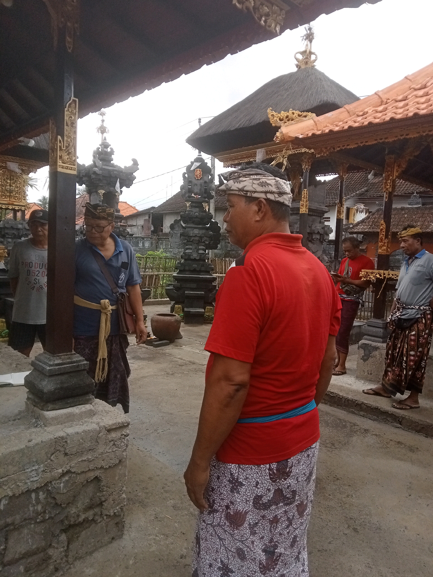 In Mendoyo, Indonesia, a group of four men stand near a Balinese temple. One man in the foreground wears a red t-shirt and a patterned sarong. To his left, another man in a blue polo shirt and a patterned sarong looks down. To the far left, a man in a gray t-shirt and dark shorts stands partially obscured by a pillar. In the background, a fourth man in a red t-shirt and dark pants is visible. The temple features traditional thatched roofs, intricate carvings, and stone altars. The ground is paved with concrete, and the sky is overcast.