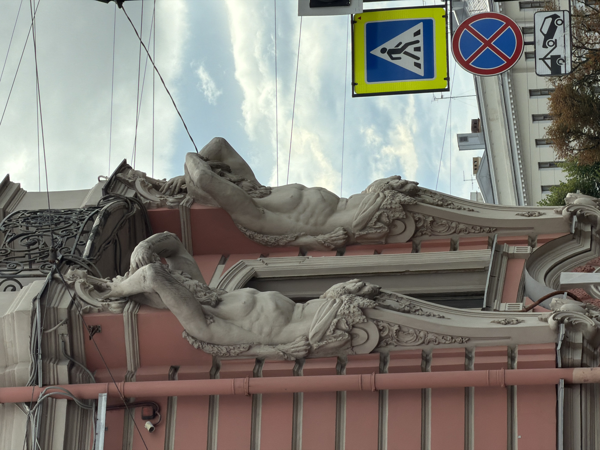 The image shows two caryatids, which are sculpted female figures used as architectural supports, adorning the facade of a building in Saint Petersburg, Russia. The caryatids are muscular and are depicted holding up a balcony. They are made of a light-colored material, likely stone or stucco, and are detailed with drapery around their lower bodies and hair. The building itself has a pink facade with decorative architectural elements. In the background, there are traffic signs including a pedestrian crossing sign and a no parking sign, along with a street with parked cars. The sky is cloudy.