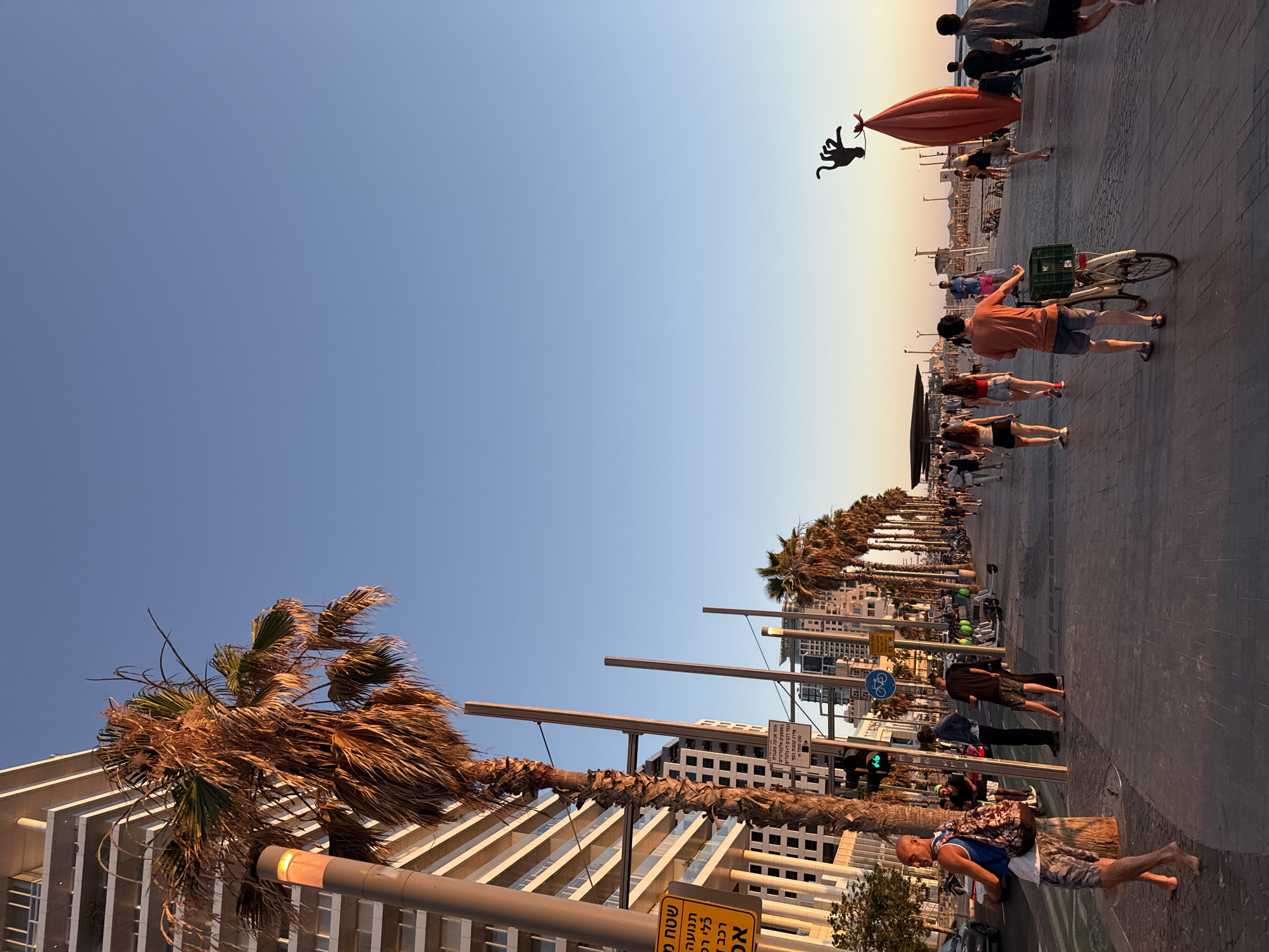 This is a wide-angle, eye-level shot of a bustling boardwalk in Tel Aviv, Israel, during what appears to be late afternoon or early evening, indicated by the warm, golden light. The boardwalk is paved with textured gray tiles and is lined with palm trees, some of which show signs of dryness. To the left, a modern, multi-story building with white balconies dominates the skyline. A lamppost with an orange glow stands near a large palm tree.
Numerous people are walking along the boardwalk in both directions, engaged in various activities. Some are strolling, while others are riding bicycles or electric scooters. In the foreground, a man with a backpack walks barefoot, carrying something in his hand. Further down the boardwalk, a man in an orange shirt pushes a bicycle with a green crate attached to the back.
In the middle ground and background, more individuals and groups are visible, some appearing to be heading towards or away from the beach. Beach chairs and umbrellas are set up in the distance. A distinctive red, elongated sculpture with a black, octopus-like figure on top stands prominently on the right side of the boardwalk, adding an artistic element to the scene.
Traffic is visible on the street adjacent to the boardwalk, with cars and motorcycles. Road signs, including traffic lights and a bicycle lane sign, are also present. The sky is a clear, soft blue, transitioning to a lighter hue near the horizon, suggesting the approaching sunset. The overall atmosphere is one of vibrant urban life and leisure.