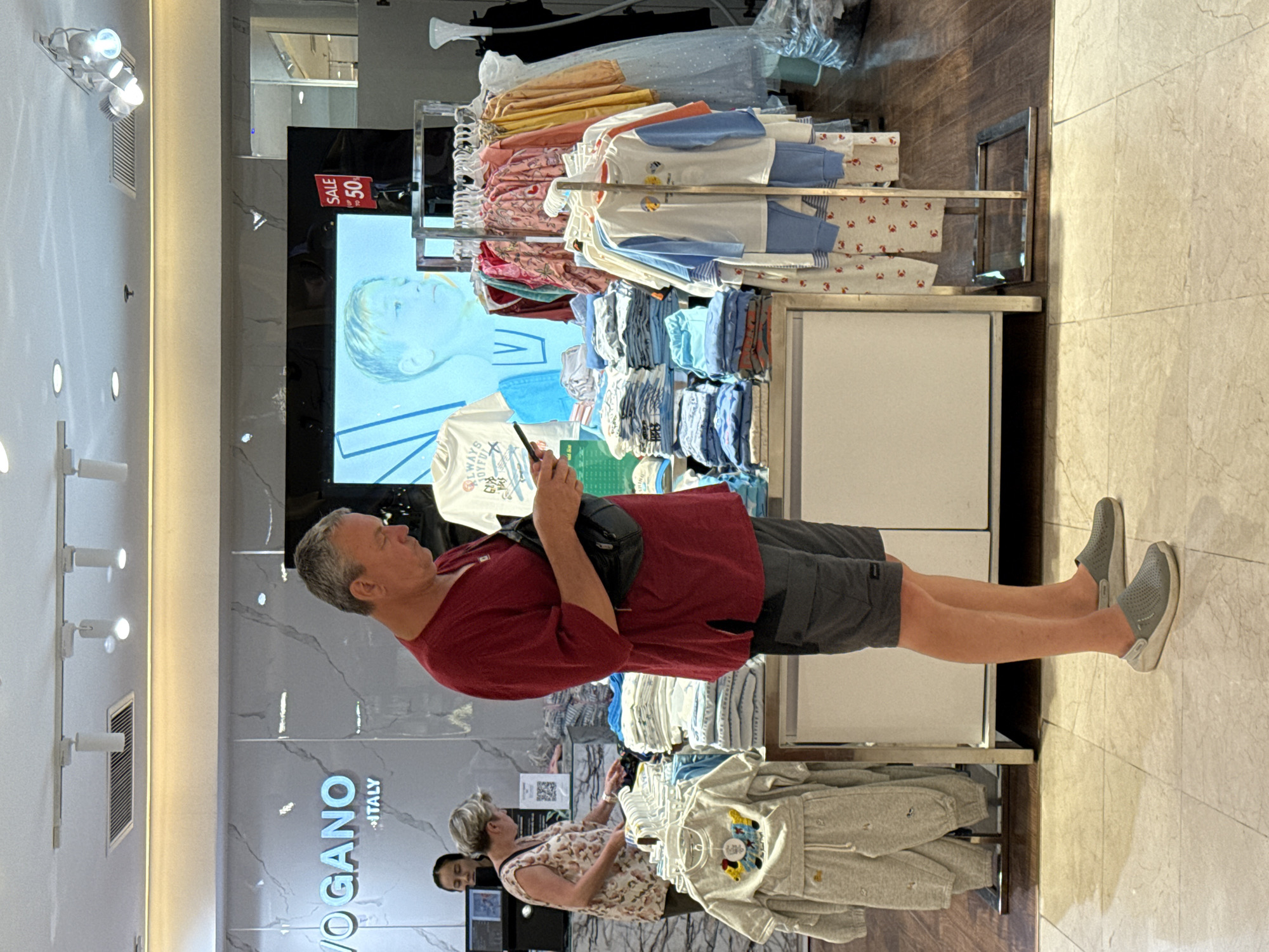 A man in a red shirt and gray shorts is standing in a clothing store, looking at his phone. He is wearing gray clogs. In the background, there are racks of children's clothing, a display screen showing a boy's face, and a sign that says "SALE 50%". Two women are browsing clothing in the background. The store appears to be a children's clothing boutique located in Nha Trang Ward, Vietnam. The store is well-lit and organized, with marble flooring and a modern aesthetic. The overall impression is one of a typical retail environment.
