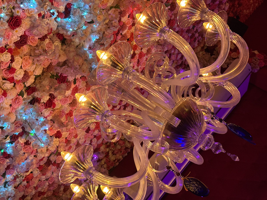 The image presents an upward-angled view of an ornate, multi-armed glass chandelier against a densely decorated ceiling. The chandelier, crafted from clear, possibly Murano-style glass, features numerous curved and spiraling arms culminating in upward-facing, fluted glass cups, each containing an illuminated candle-style bulb emitting a warm, yellowish light. Darker, possibly metallic or deep amber glass elements form the central structure, from which hang several dark blue or black teardrop-shaped glass pendants on beaded strands. The ceiling directly above and around the chandelier is entirely covered with a dense arrangement of artificial flowers, predominantly roses, in shades of light pink, dark pink, red, and cream. Small, bright blue string lights are interspersed within this floral canopy. The overall lighting is dim, with the chandelier and blue lights providing the primary illumination. A muted red surface is visible in the lower corners, and a white air conditioning vent is partially obscured below the chandelier. No individuals are visible. This decorative style is characteristic of upscale cafes, restaurants, or hotel interiors commonly found in Paris, France, known for their opulent and aesthetically curated environments.