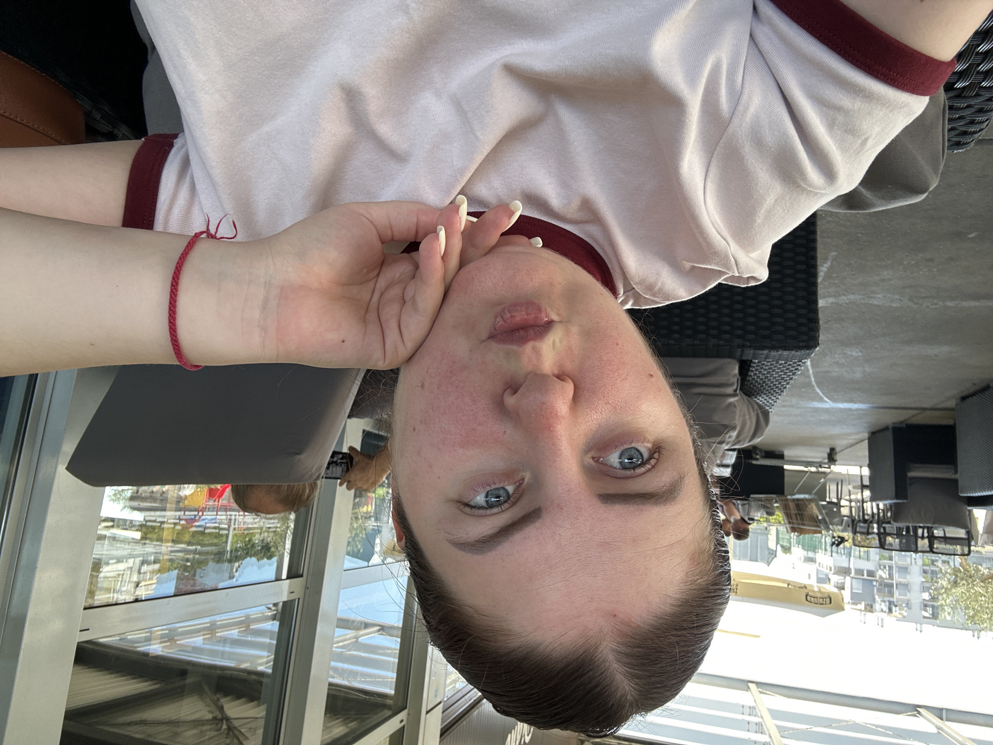 In the city of Grad Rijeka, Croatia, a young Caucasian woman with blue eyes and brown hair pulled back is captured in a close-up selfie. She is wearing a light pink t-shirt with a maroon trim around the neckline and sleeves. Her right hand, with white-nailed fingers, rests on her chin, and she has a red braided bracelet on her left wrist. The background reveals an outdoor seating area, possibly a cafe or restaurant, with wicker furniture, tables, and chairs. In the distance, a distinctive yellow and white zeppelin is visible, along with modern buildings and a playground. The scene is illuminated by natural daylight.