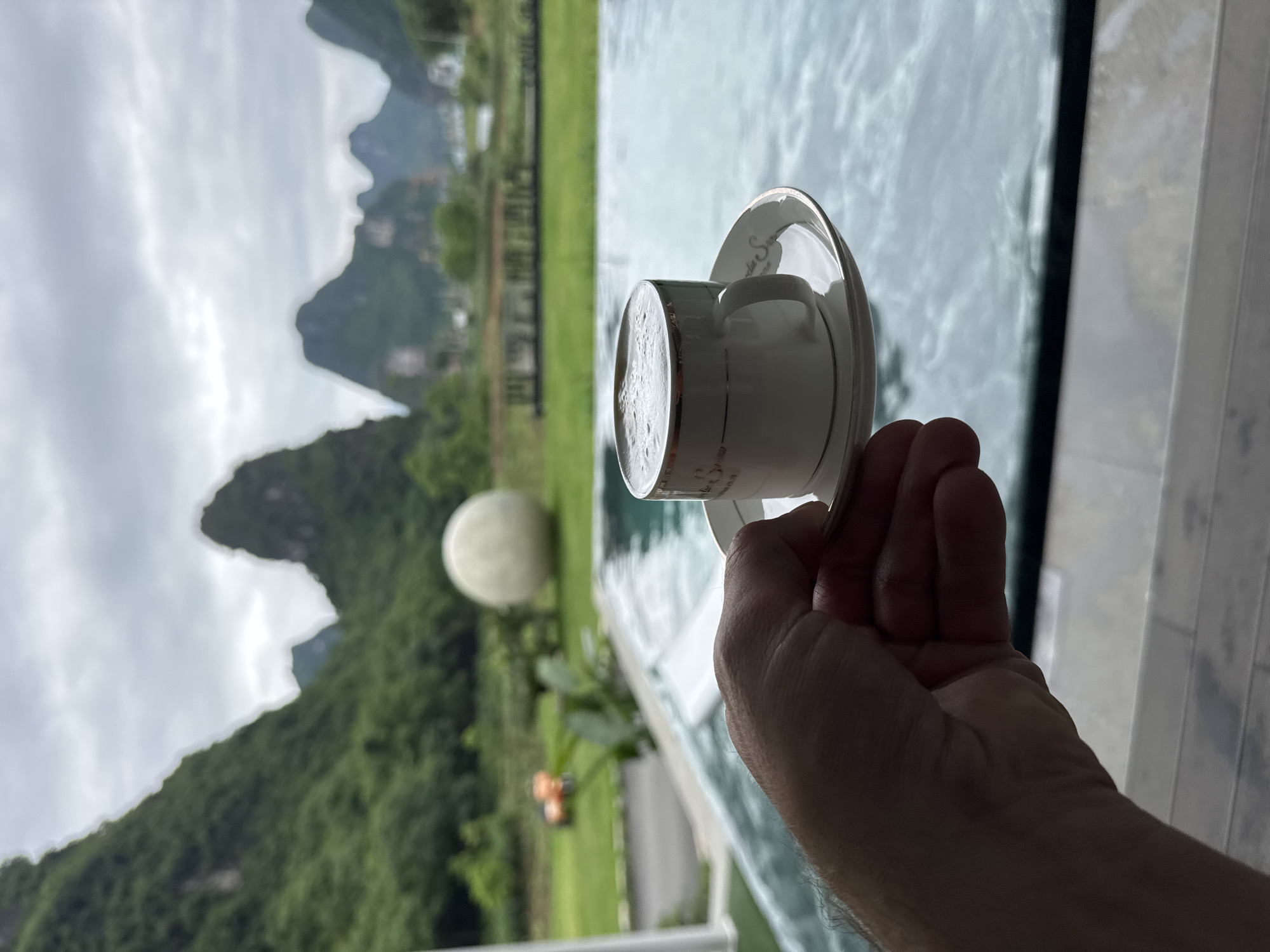 A right hand holds a white porcelain cup and saucer, filled with a foamy beverage, in front of a landscape. The landscape includes green, mountainous terrain, a white spherical object, manicured green grass, and a blue body of water. The scene appears to be in Guilin, China, based on the karst topography of the mountains. The hand and cup are situated near a white railing. The sky is overcast. The image appears to have been rotated clockwise.