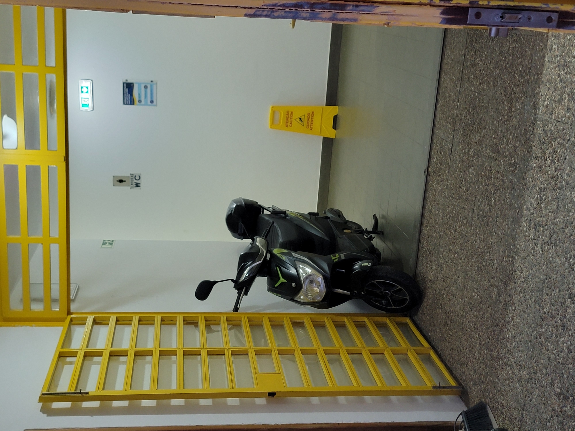 An open, yellow metal gate with a lattice design reveals an interior space. A black and green electric scooter is parked just inside the gate. The scooter has a black helmet resting on its rear seat. In the background, on a white wall, are two signs. The left sign depicts a stick figure in a circle with the text "WC" below it. To the right, an illuminated exit sign with a green arrow and a figure is mounted high on the wall. Further to the right, a yellow "Wet Floor" cautionary sign is propped up. The floor in the interior is tiled. The overall environment suggests an entrance to a building, possibly a public facility or residential complex in Quarteira, Portugal, given the context.