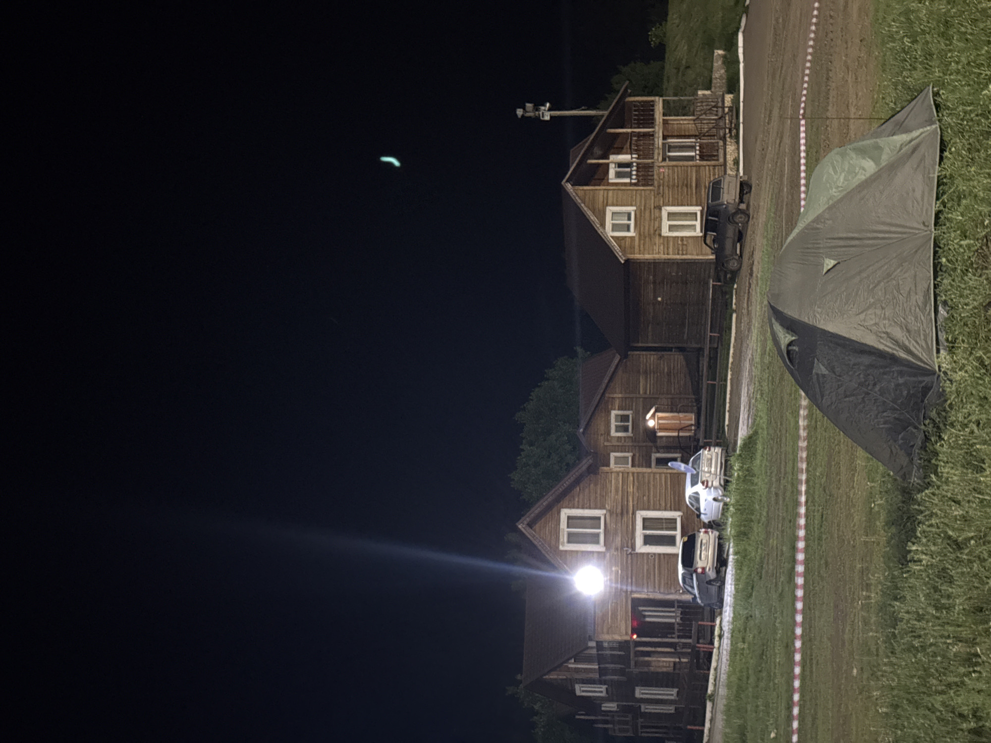At night in Khvalynsk, Russia, several multi-storied wooden buildings stand adjacent to a grassy area. A dark-colored tent is pitched on the grass. A red and white barrier tape runs alongside the grassy area, near several parked vehicles. A bright light illuminates one of the buildings. The sky is dark with a streak of light.