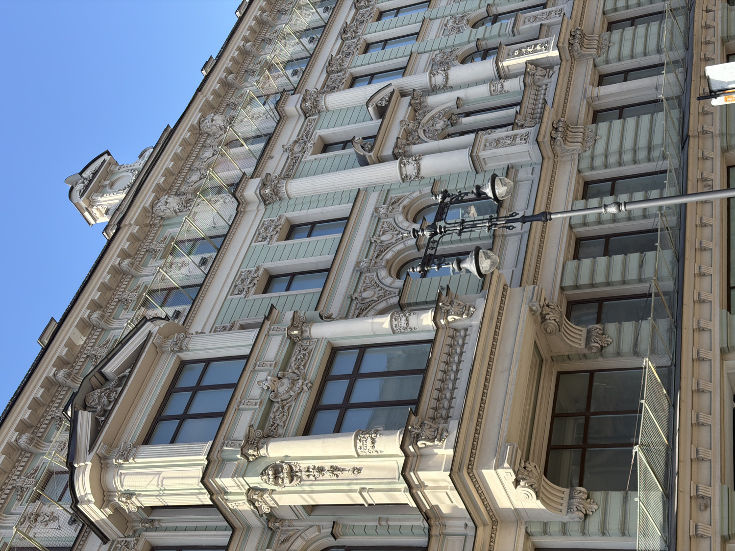 The image presents a low-angle, upward view of the highly ornate facade of a historic building, likely situated in Saint Petersburg, Russia, under a clear blue sky. The multi-story structure is distinguished by its intricate architectural details, predominantly in an off-white or cream color, with accents of pale green or blue on recessed panels between the windows.
The facade is rich with decorative elements characteristic of classical or Baroque revival styles, including fluted columns, ornate capitals, elaborate carvings such as festoons, volutes, and corbels, as well as decorative moldings and cornices. Numerous rectangular windows with dark frames are visible, many set within deep recesses. Some windows appear to have a fine mesh or safety netting extending from them, visible particularly on the right side.
A prominent, dark-colored lamppost with two antique-style luminaires extends diagonally across the mid-ground, further emphasizing the urban setting. The bright, strong sunlight illuminates the facade from the left, creating deep shadows that highlight the three-dimensional relief and texture of the architectural decorations. The sky in the upper left corner is a brilliant, cloudless blue.
No people or discernible text are visible in the image. The scene conveys a sense of grandeur and meticulous craftsmanship, reflecting the historical architectural heritage of the likely location.