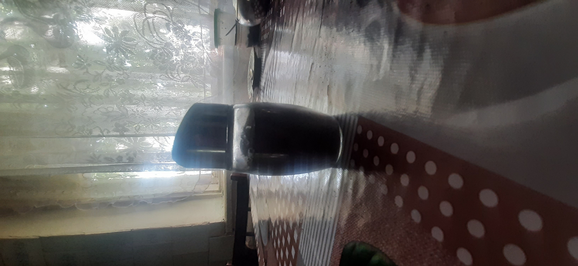 A glass jar with a black lid, filled with a dark substance, sits on a patterned tablecloth in front of a window with lace curtains. The jar is in the center of the image, slightly to the right. The tablecloth has a repeating pattern of white dots on a reddish-brown background, and a section with gray and white stripes. Behind the jar, sunlight streams through the sheer lace curtains, illuminating the room. The overall impression is of an indoor domestic setting, likely a kitchen or dining area, in Yessentukskaya, Russia. There is no indication of any specific time or action.