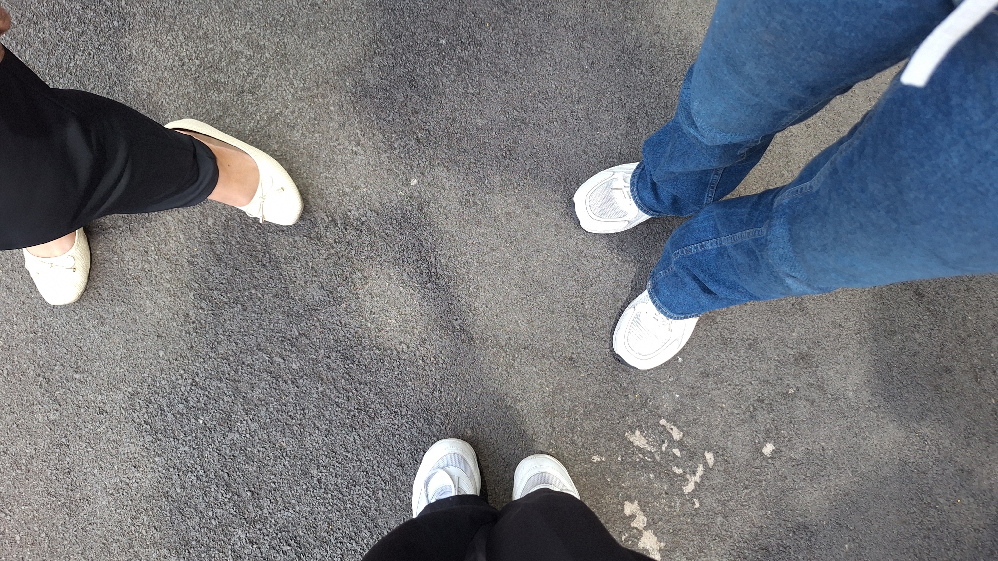 The image presents a high-angle, close-up view of the lower legs and feet of three individuals standing on a dark grey, pebbled asphalt surface. The setting appears to be outdoors, likely on a paved road, pathway, or in a parking area in Karawang, Indonesia.
On the left, one person wears black trousers and light-colored, possibly cream or off-white, ballet flats or slip-on shoes featuring a small bow detail. Another person is positioned to the right, dressed in blue denim jeans and white athletic sneakers with mesh elements. The third individual is seen at the bottom of the frame, wearing dark trousers (likely black, similar to the person on the left) and white athletic sneakers.
The three people are standing in a loose triangular formation, suggesting they might be paused, conversing, or simply gathered. The activity is static, with no dynamic motion captured. The lighting indicates it is daytime, casting soft or no discernible shadows. There is no visible text on any of the clothing, shoes, or the ground, and due to the cropping, no emotions can be interpreted.
