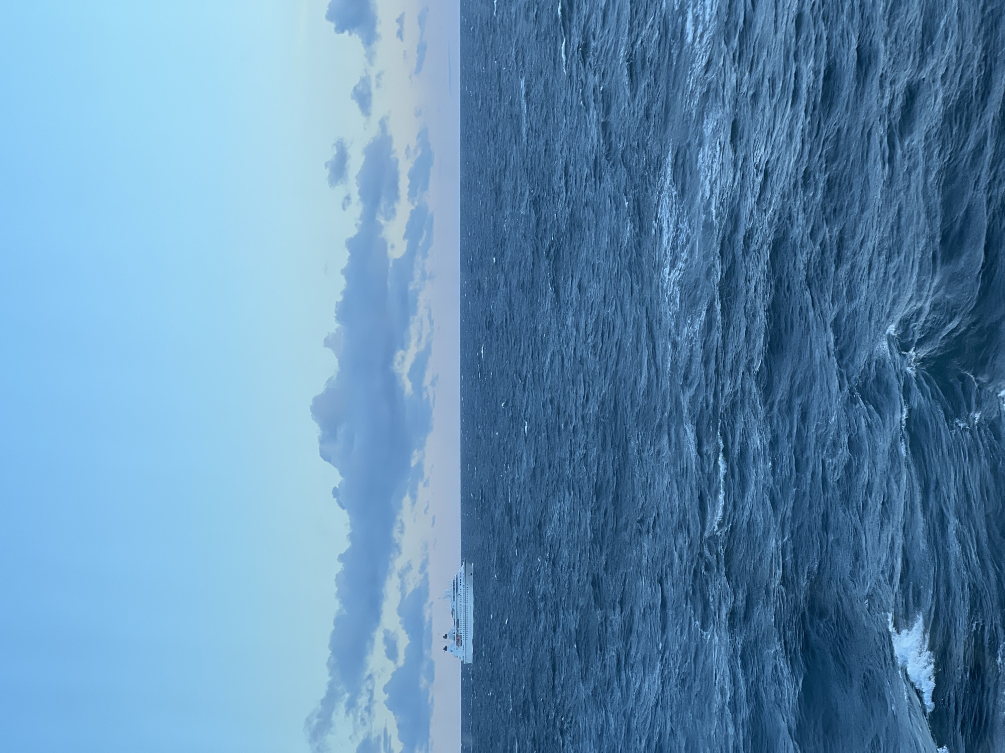 A wide shot captures a cruise ship sailing on choppy, dark blue ocean water under a clear, pale blue sky with scattered grey clouds near the horizon. The ship, painted white with black accents, is positioned in the upper left quadrant of the frame. The water exhibits a textured surface with visible waves and whitecaps, suggesting moderate movement. The horizon line is sharp and distinct, separating the sea from the sky. The overall atmosphere is serene and vast, with a cool color palette dominated by blues. There is no visible indication of the location being in a city in Poland.
