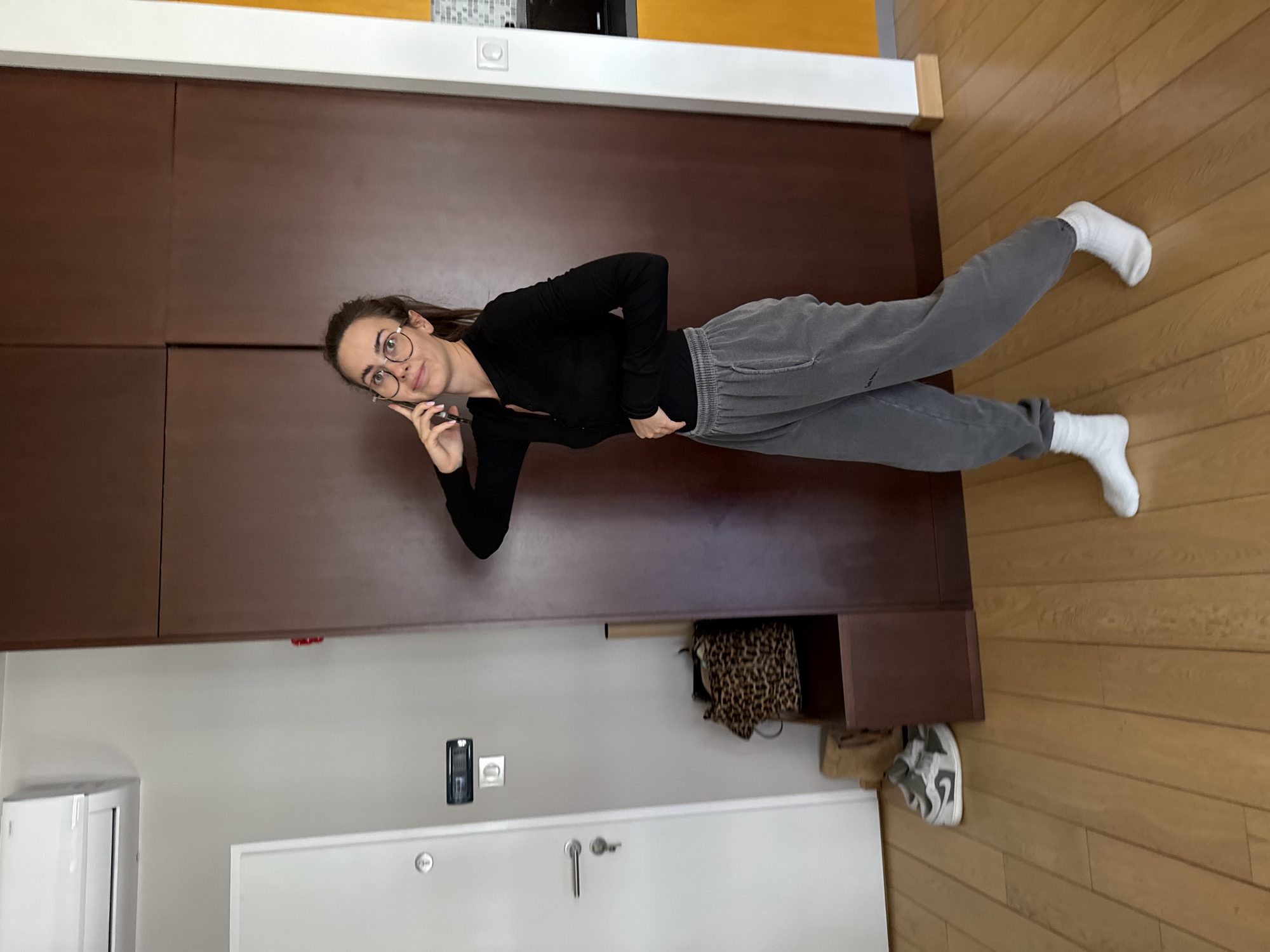 The image displays a young woman standing casually in an indoor residential setting, likely in Warsaw, Poland. She is positioned against a large, dark brown wooden cabinet or wardrobe that takes up a significant portion of the background. The woman has dark hair, wears round-framed glasses, a long-sleeved black top, and grey sweatpants with white socks. She looks towards the camera with a subtle smile, holding a dark smartphone to her chin with one hand while the other rests on her hip.
The floor beneath her is made of light-colored wooden planks. On the opposite side of the room, a light grey wall is visible, featuring a white door with a silver handle and keyhole, and a white air conditioning unit or similar appliance. A black wall-mounted device (possibly an intercom or thermostat) and a white electrical outlet are also present on the grey wall. Within an open section of the brown cabinet, a leopard print fabric item, likely a bag, is stored alongside another neutral-colored bag. A grey and white sneaker is visible on the wooden floor near the cabinet.
The scene suggests a relaxed atmosphere, with the woman posing for the photograph. The time of day and weather are not discernible, as the lighting appears to be internal. No legible text is visible on any objects or surfaces within the frame.