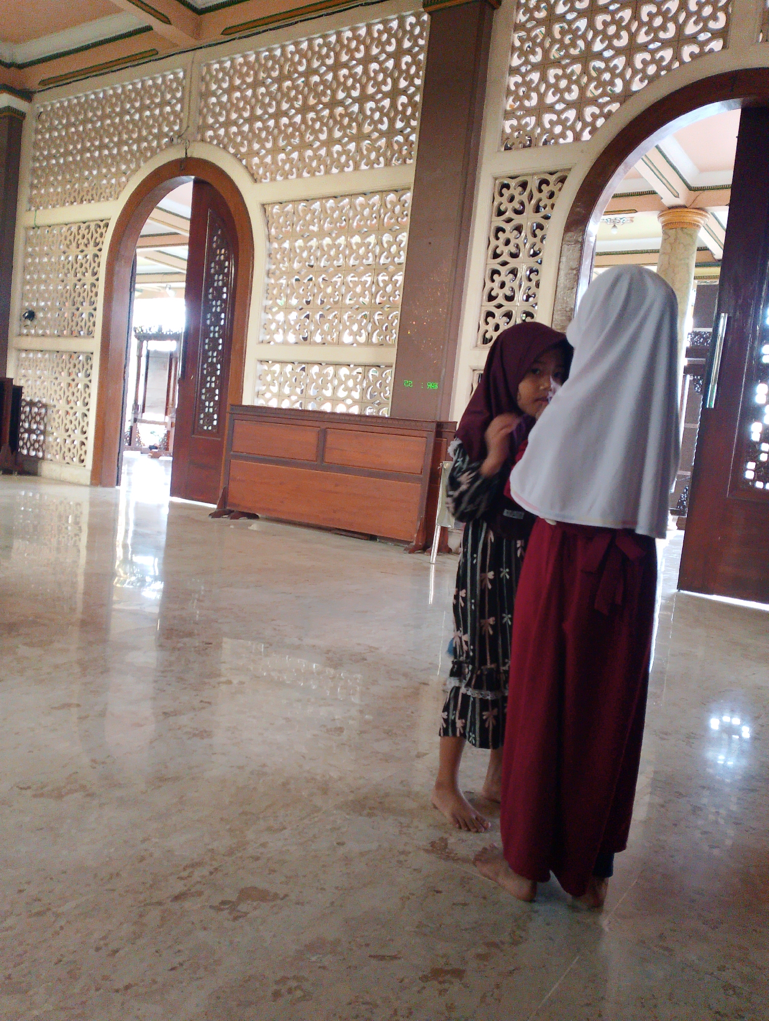In a sunlit interior, possibly a mosque due to the ornate latticework on the walls and the presence of two girls in hijabs, a young girl wearing a dark patterned dress and a maroon hijab stands to the left, looking towards the viewer. To her right, another girl in a white hijab and a long red skirt stands with her back mostly to the camera. They appear to be conversing in a tranquil setting with a polished, reflective floor. The architectural details suggest a building with traditional Indonesian influences, located in Sukodono, Indonesia.