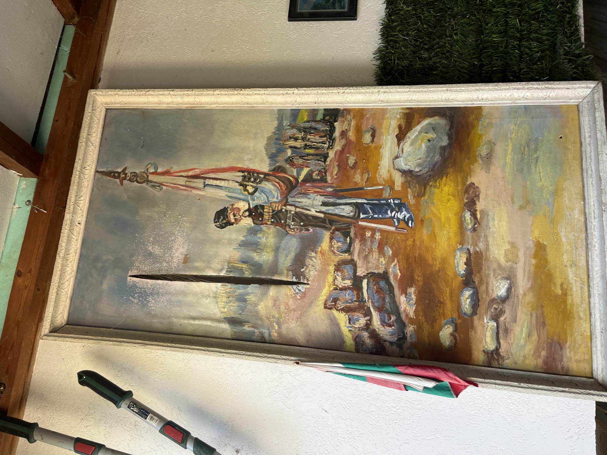 The image shows a framed painting hanging on a wall in an indoor environment. The painting depicts a soldier with a flag in a landscape with other figures. The background consists of a sky and land with indistinct features. The painting is in a white ornate frame. Other details visible include part of a plant, a section of what looks like a wooden structure, a dark-colored framed picture above the painting, a section of a pole with red/green highlights, and hedge trimmers along the lower left portion of the picture. The location is indicated as Unknown, Unknown.