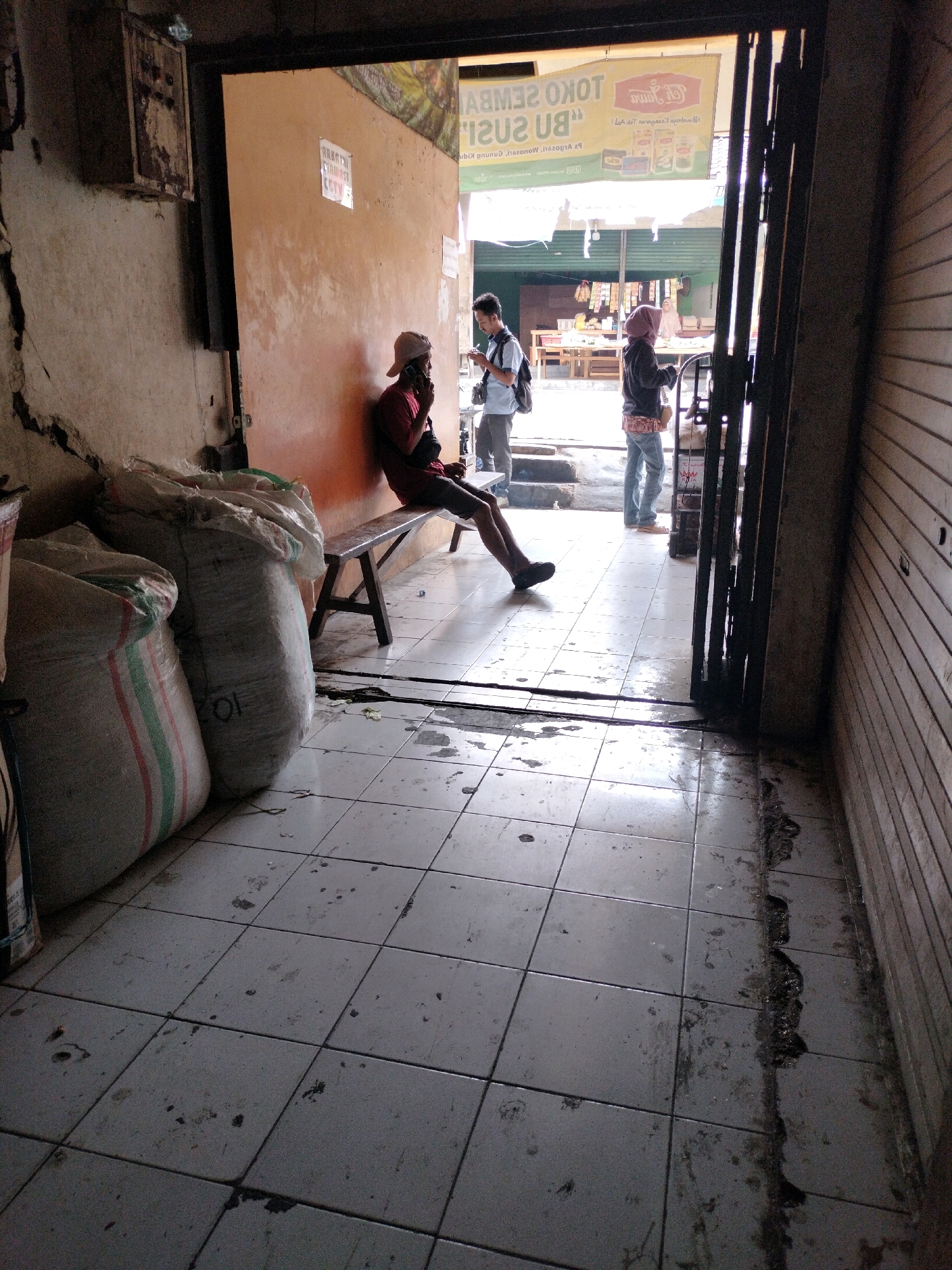 The image shows a covered outdoor area with tiled flooring. In the foreground, two large sacks, possibly containing goods, are stacked. A person is seated on a wooden bench to the left, leaning against an orange-colored wall, wearing a red shirt and a white cap, and appears to be talking on a mobile phone. Further into the background, two other individuals are standing. One, a male wearing a white shirt and a backpack, is looking down at a phone. The second, a female wearing a dark top and jeans, stands facing away from the camera. The background reveals an open market area with stalls and more people. The scene appears to be located in a semi-urban or market setting, consistent with locations in Wonosari, Indonesia, given the architectural style and general ambiance. The floor is tiled and appears wet and somewhat dirty, suggesting recent rain or cleaning. The overall impression is of a bustling but somewhat utilitarian commercial space.