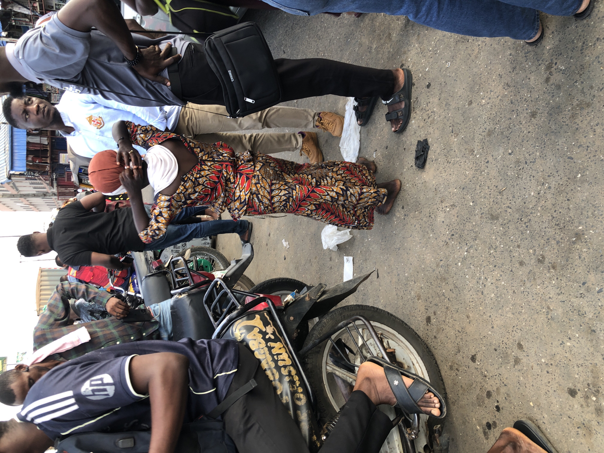 The image depicts a street scene in Ikeja, Nigeria. In the foreground, a child wearing a vibrantly patterned dress with red, yellow, and black designs is standing with their back to the camera, adjusting a headscarf or head covering. To the left, a person on a motorcycle is partially visible, wearing a dark blue shirt with white stripes and an HP logo. The motorcycle has a black fender with gold lettering that appears to read "OMAGBA" and other text. To the right of the child, a man in a light blue collared shirt and beige pants is looking to the left. Next to him, another man in a grey short-sleeved shirt and black pants is facing away, with a black messenger bag slung across his chest. The ground is asphalt, with scattered debris including plastic bags. In the background, other people, motorcycles, and a building with a blue awning are visible, suggesting a busy urban environment. The lighting suggests it is daytime.