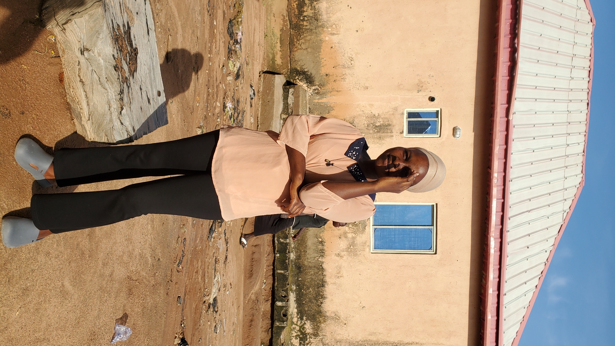 A young woman stands facing the camera in front of a peach-colored building with a corrugated metal roof. She is wearing a light beige headscarf, a peach-colored blouse with a dark, beaded collar, black pants, and grey clogs. Her right hand is raised to her forehead, obscuring her eye. The building has two rectangular windows with blue-tinted glass. The ground is sandy and uneven, with scattered debris. A large, weathered wooden beam lies in the foreground to the left, casting a shadow. Another person, partially visible, walks behind the woman in the background. The scene appears to be outdoors in a casual, possibly semi-urban or rural, setting in Jalingo, Nigeria.