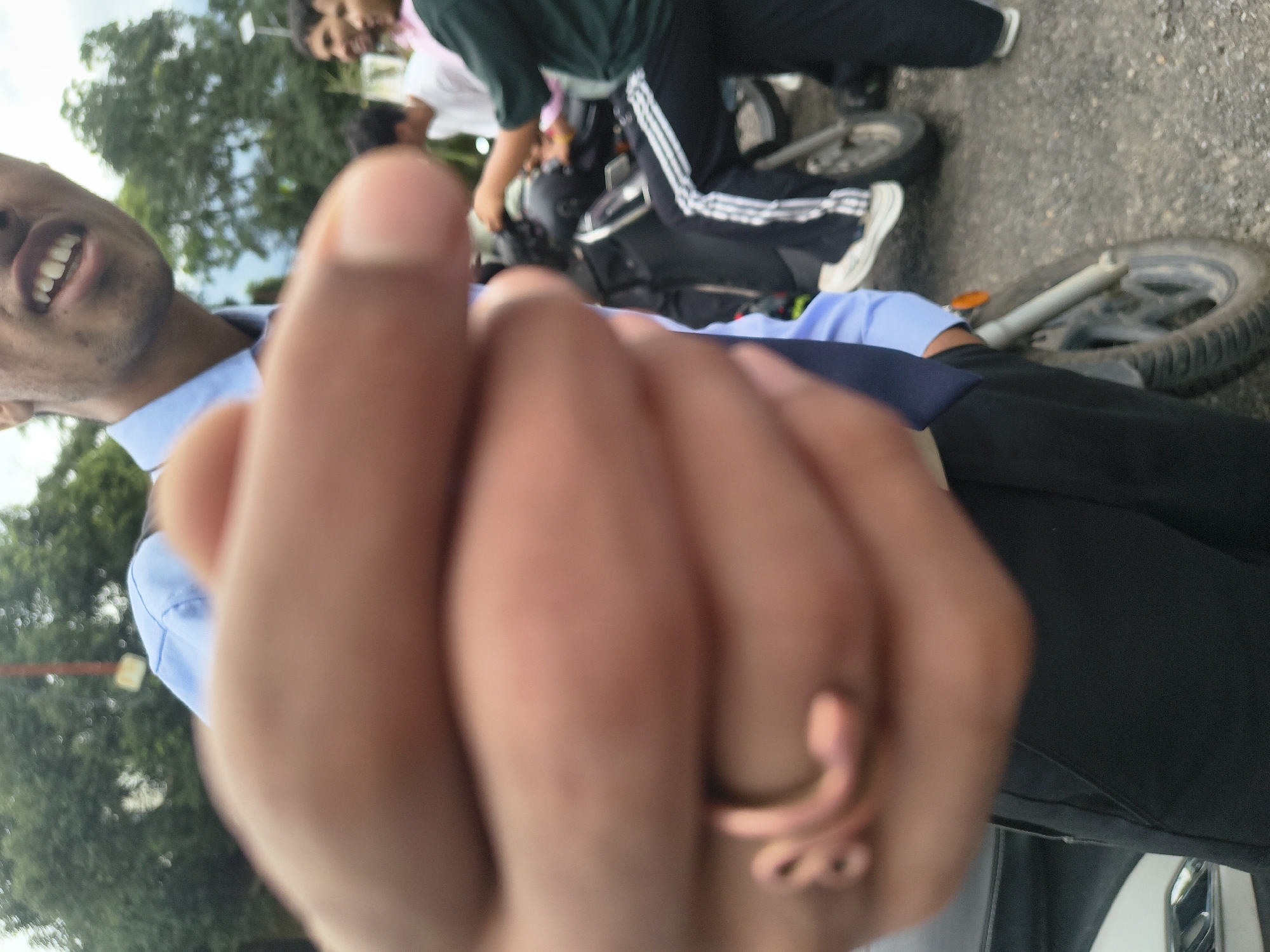 An out-of-focus, close-up shot shows a person's hand with a ring in the foreground, obscuring the face of the individual. The person appears to be wearing a light blue collared shirt and dark trousers. In the background, two other individuals are visible, one wearing a green shirt and dark pants with white stripes, and another in a white t-shirt. A motorcycle is parked in the mid-ground, and the overall setting suggests an outdoor urban environment, possibly in Dharamshala, India, given the presence of trees and what appears to be a road or paved area. The light is somewhat overcast.