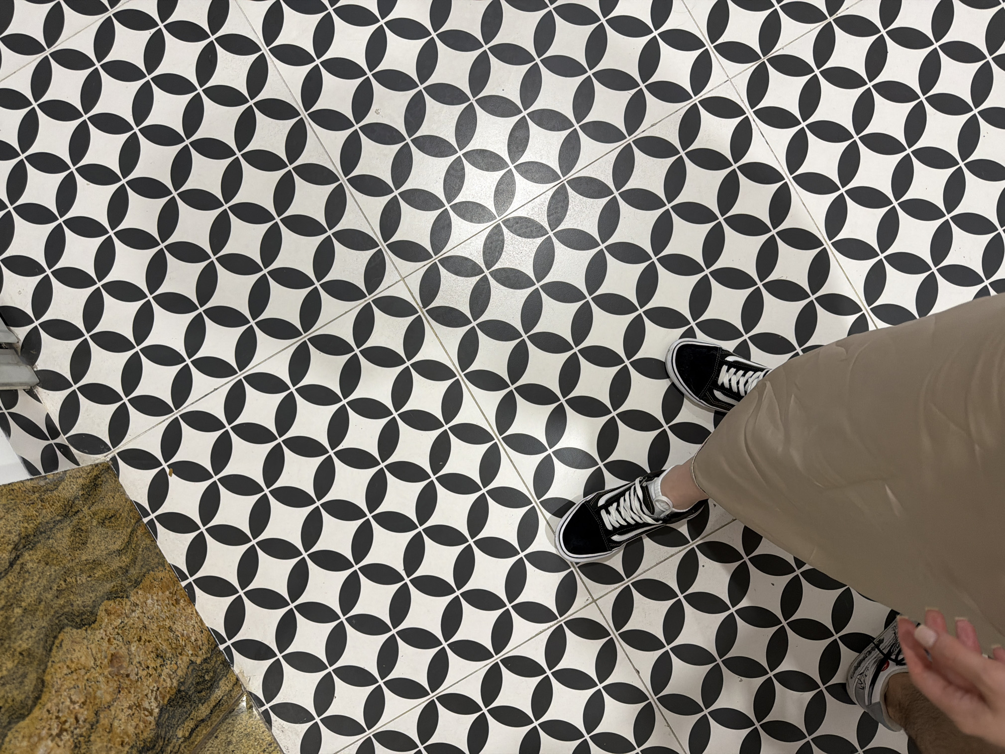 A high-angle shot shows a person standing on a black and white patterned tile floor. The person is wearing black and white sneakers, a light brown skirt, and white socks. The floor pattern is a repeating design of overlapping black circles on a white background. Part of a different type of flooring, possibly stone or marble, is visible at the bottom left corner. A glimpse of the wall or adjacent surface is seen in the upper left corner, suggesting this is an indoor setting. The image provides no information to indicate this scene is located in Đà Nẵng, Vietnam.