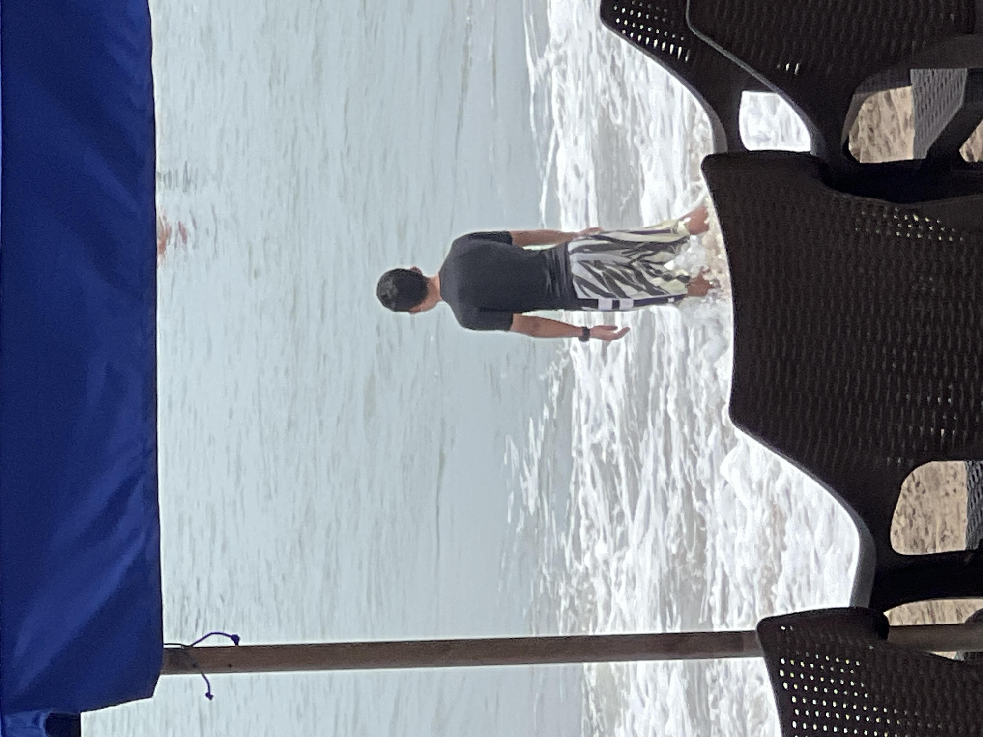 A young man stands with his back to the camera, facing the ocean on the shore of Kuta Beach, Indonesia. He is wearing a black short-sleeved rash guard and striped swim shorts. The waves are reaching his shins. In the foreground, two dark brown plastic beach chairs are visible, positioned on the sand. Above, a blue canopy shades the area. The sky is overcast, and the water appears a muted grey-blue.
