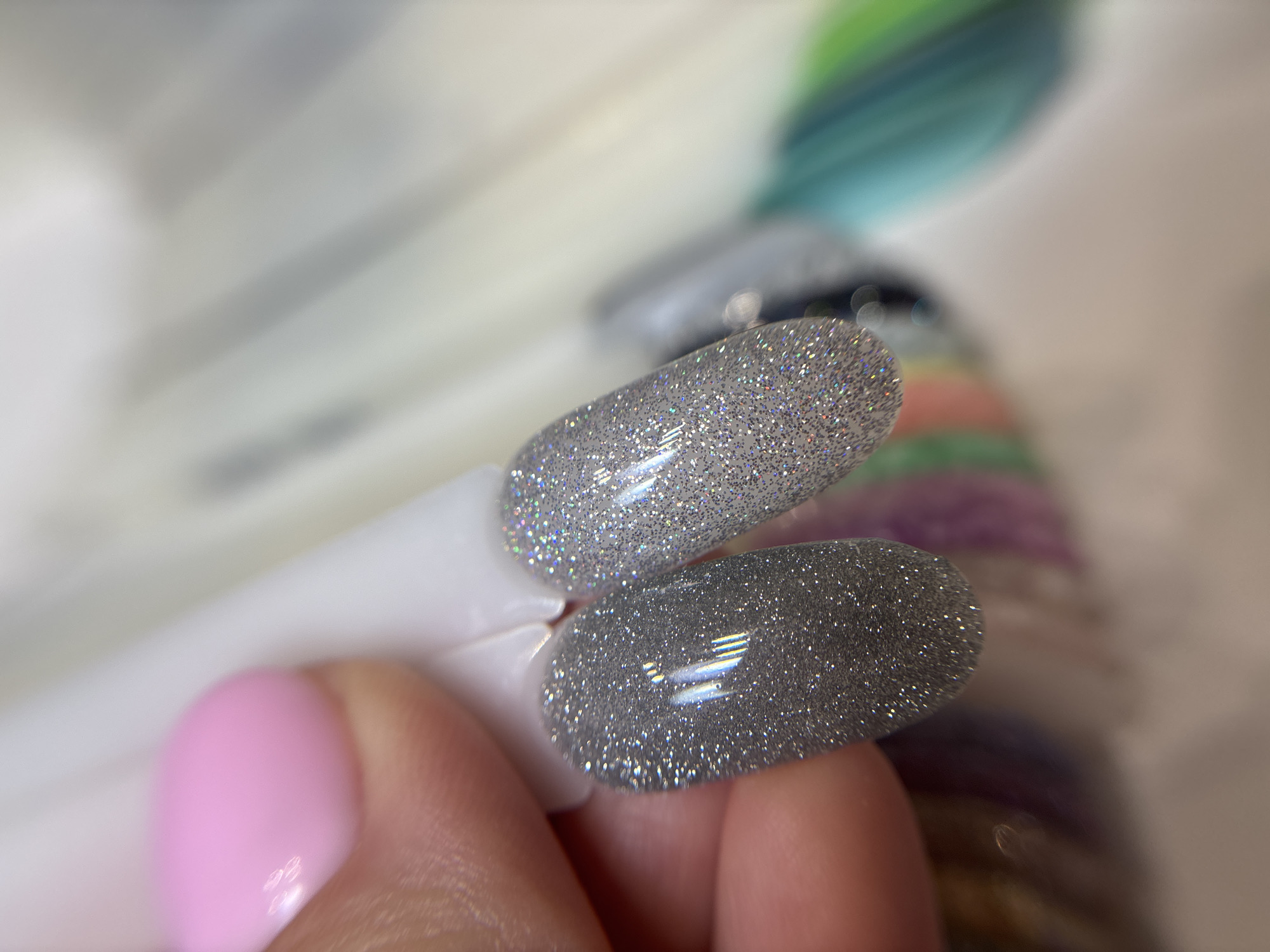 A human hand, featuring a manicured thumb with light pink polish, holds two artificial nail tips for display. The nail tip on the left is an opaque dark silver with concentrated glitter particles, exhibiting strong specular reflections from an overhead light source. The nail tip on the right is an opaque light silver with similar glitter application and reflections, appearing brighter than its counterpart. Both tips have an almond or oval shape. In the blurred background, an array of other colored nail swatches on similar white plastic bases is visible, suggesting a nail salon or beauty supply retail environment in Ферма, Russia, where clients select nail polish colors.