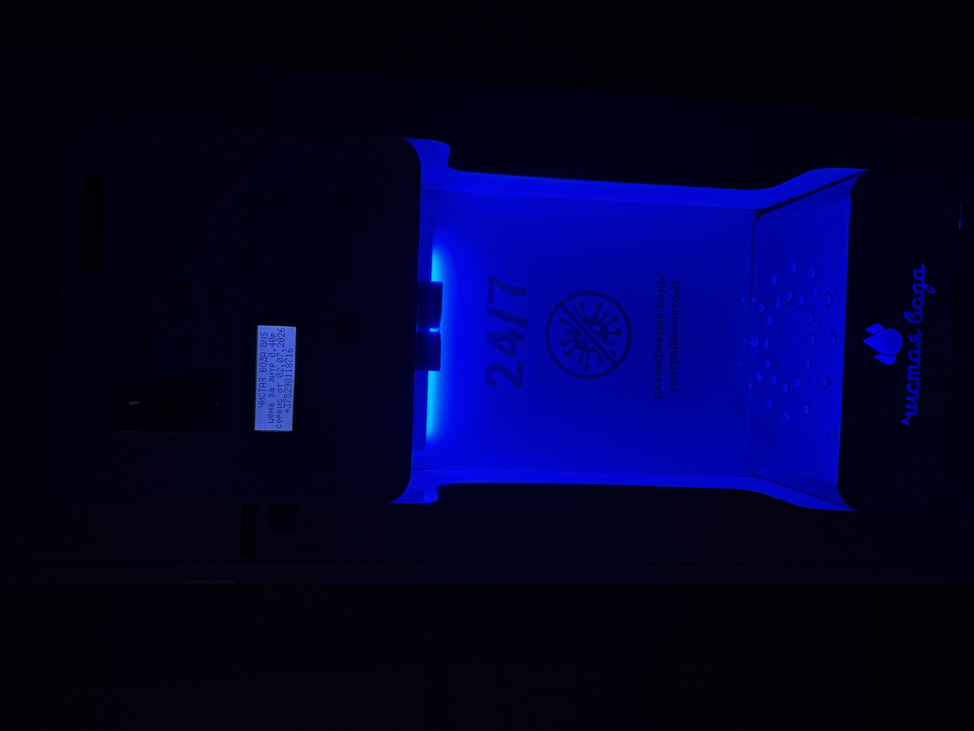 The image shows a water dispensing machine with blue lighting. On a small screen, text is displayed in Cyrillic: "ЧИСТАЯ ВОДА GNS" (Clean Water GNS), "ЦЕНА ЗА ЛИТР 0.40Р" (Price per liter 0.40R), "02.07.2026", and "+375298118716". Below the screen, the numbers "24/7" are prominently displayed in large font, along with a circular icon depicting a stylized virus crossed out, indicating disinfection. Further text below the icon reads "дезинфекция воды ультрафиолетом!" (water disinfection with ultraviolet!). At the bottom of the machine, the words "Чистая Вода" (Clean Water) are written. The machine appears to be located in an indoor, dimly lit environment. The phone number includes the Belarusian country code +375. Based on the country code and the Cyrillic text, the machine is likely located in Belarus, possibly in Vitsebsk. There are no people visible in the image.