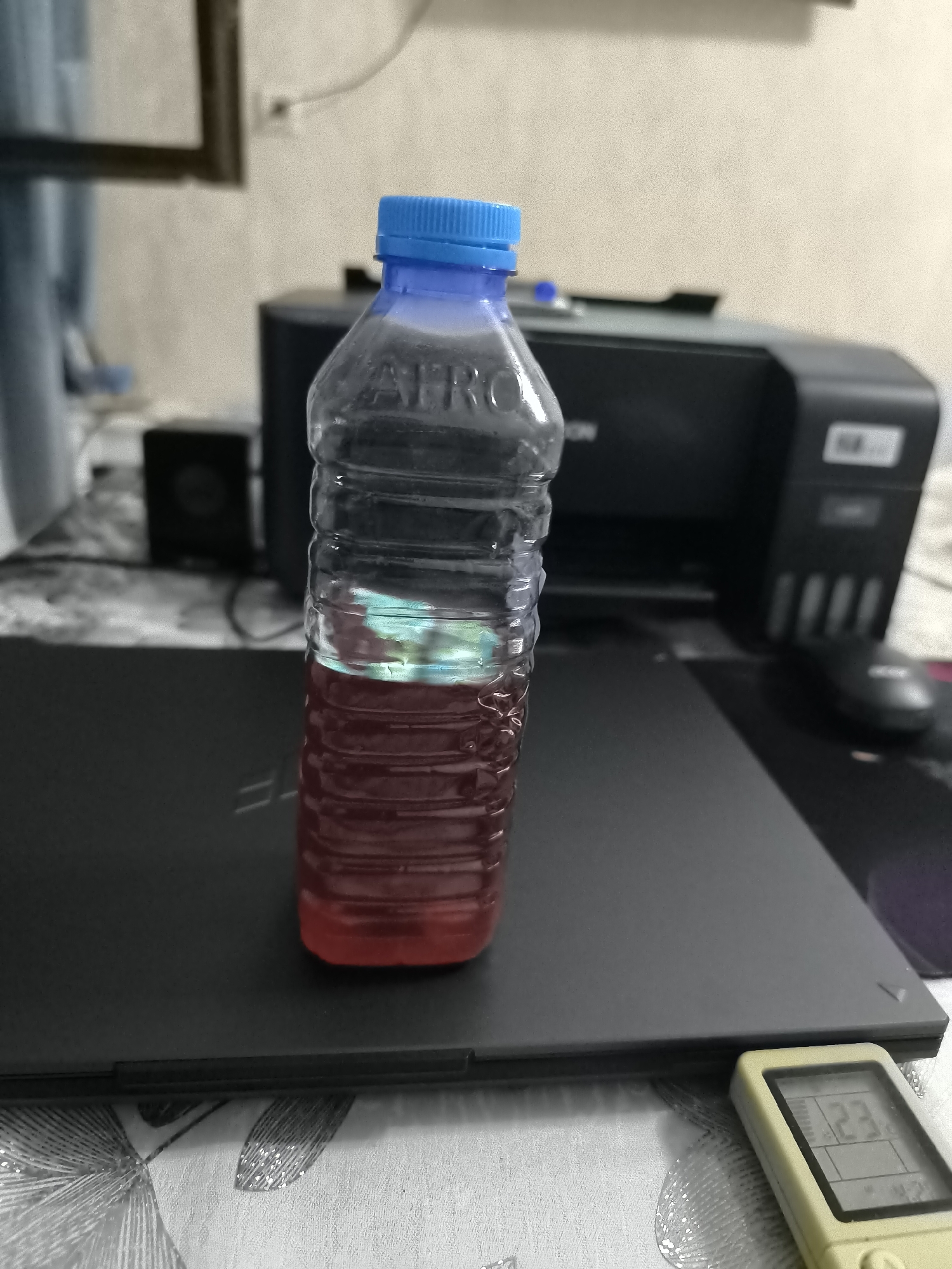 An indoor, eye-level, medium shot captures a partially filled plastic bottle with a blue cap. The bottle contains a red liquid with a layer of lighter colored liquid on top. It is placed on a dark laptop, which is positioned on a table covered with a patterned tablecloth. In the background, a black printer is visible, along with a small black speaker. The scene is well-lit, with shadows cast by the objects. There are no people present in the image. The location is not discernible within the image, and therefore its presence in Kokand, Uzbekistan, cannot be confirmed.