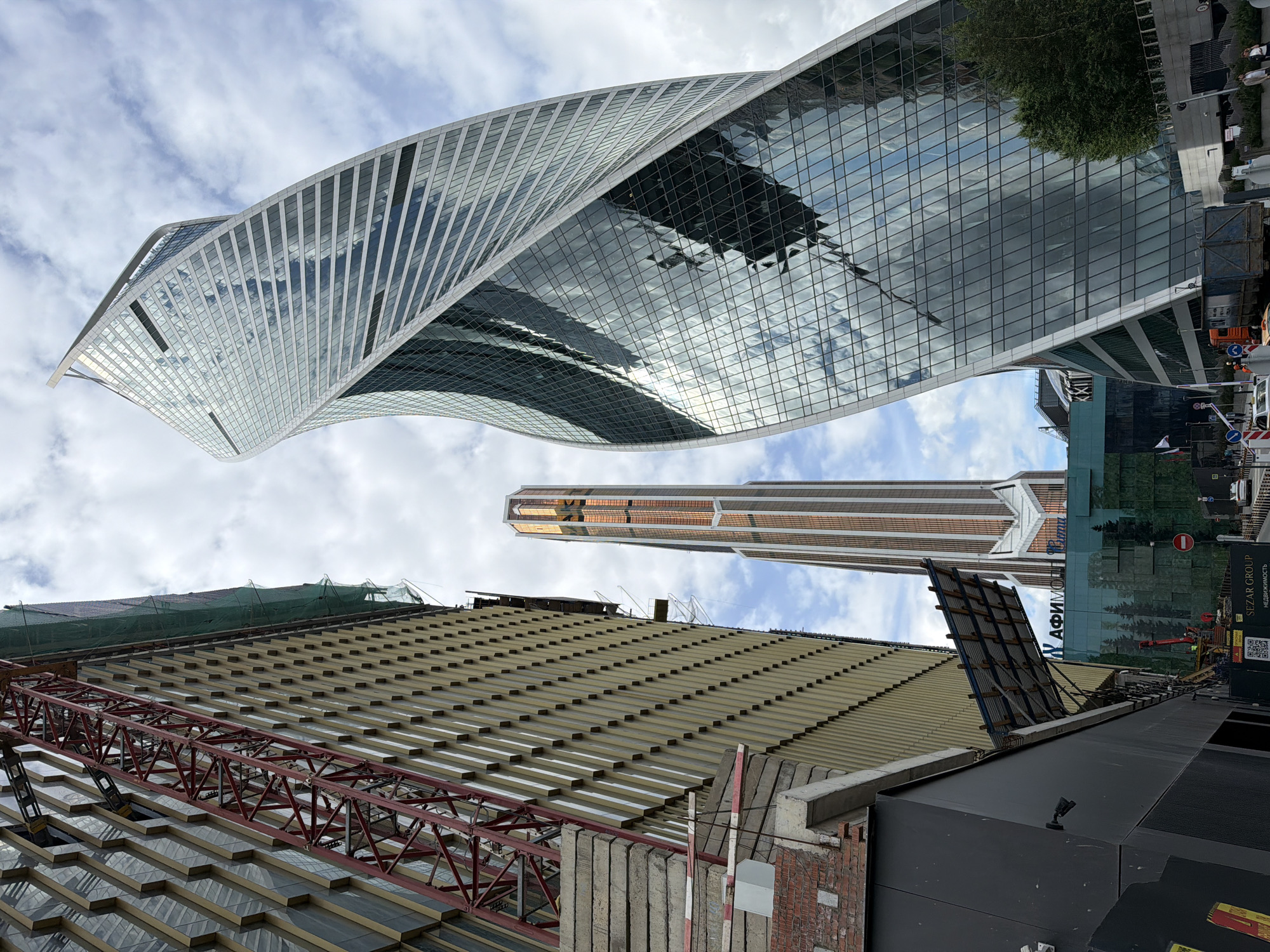 The image shows a low-angle view of the Moscow International Business Center (Moscow City), Russia. The Evolution Tower, a twisting skyscraper with a glass facade reflecting the sky and other buildings, dominates the upper half of the frame. Below it, another skyscraper is visible, with a copper-colored vertical stripe design. The lower half of the image includes a building under construction, indicated by a red metal crane structure, exposed brick, and other construction materials. A large section of a building in the bottom right with advertising graphics on a construction screen is also seen. The sky is overcast with scattered clouds.