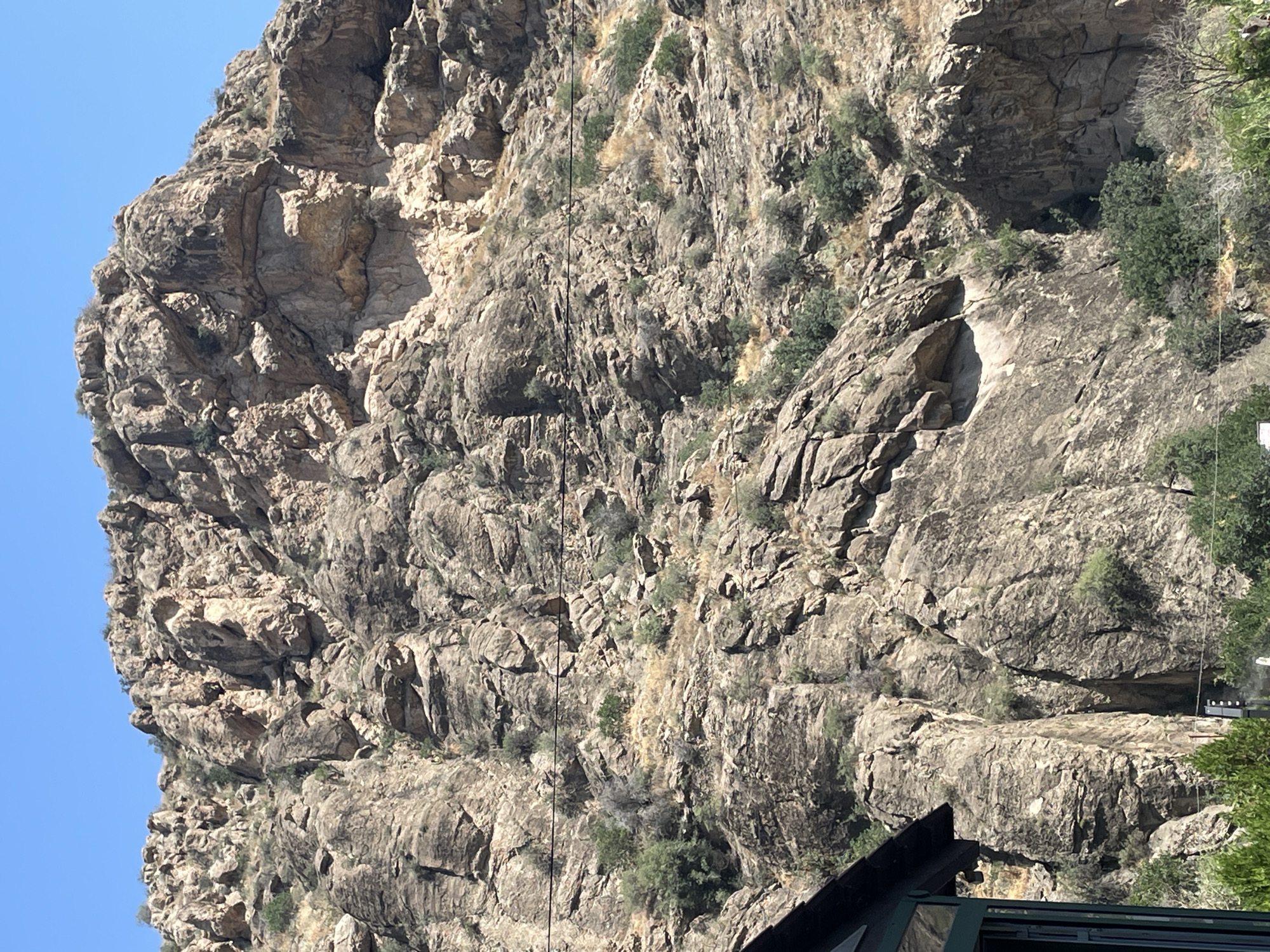 This is a photograph taken in Areni, Armenia, featuring a rugged, rocky mountainside under a clear blue sky. The mountain is predominantly composed of grey and brown rock formations, with visible cracks, crevices, and uneven surfaces. Sparse vegetation, including dry grasses and small green bushes, dots the mountainside, particularly in the lower and middle sections.
In the bottom left corner of the image, a portion of a dark-colored structure with a green-tinted window frame is visible, suggesting the presence of a building or shelter. Two thin, dark horizontal lines, likely power lines, stretch across the mid-section of the mountainside. The overall environment appears arid and mountainous. There are no people or animals visible in the frame.