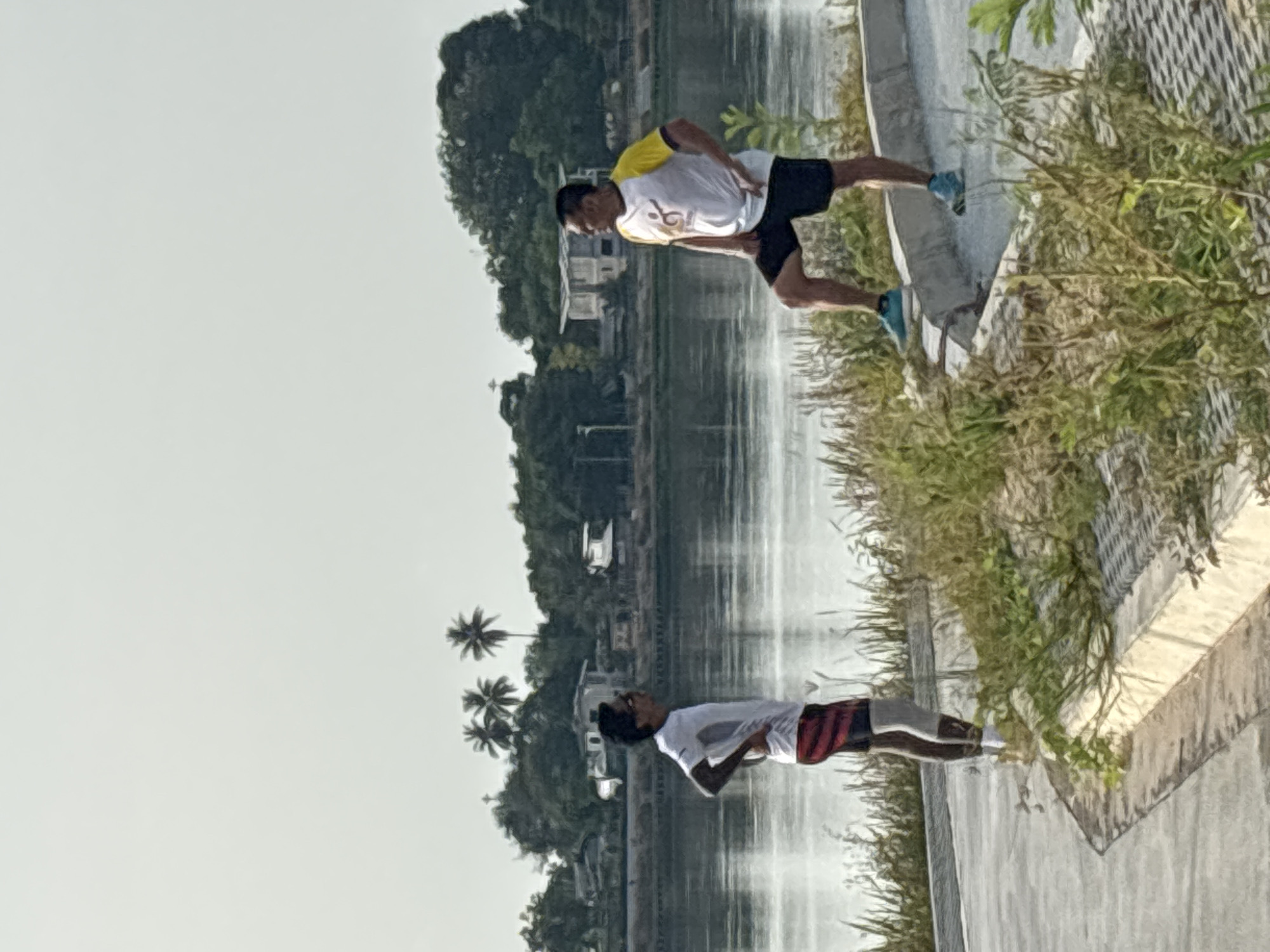 Two men are pictured near a body of water in บ้านบางรักษ์, Thailand. One man, wearing a white shirt with yellow sleeves and black shorts, is running. The other man, wearing a white shirt and striped shorts, is standing with his arms crossed. The scene includes trees, buildings, and a concrete embankment.