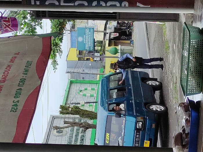 The image captures a daytime street scene in Kepanjen, Malang Regency, Indonesia, viewed from an indoor vantage point, likely a shop or building. In the foreground, a large banner with red and white sections is partially visible on the left, displaying reversed text "RAZOLOM AYAR" and a phone number "TELEPON: 0851 4103 5422". On the right, a green plastic laundry basket sits on what appears to be a counter or floor.
The central activity involves a man standing outside a blue Mitsubishi L300 van, interacting with the driver. The man outside wears a light blue long-sleeved shirt, a dark vest, dark trousers, and a wide-brimmed hat. He is leaning towards the open window of the van, appearing to converse with the driver. The driver, wearing a white shirt, is visible in the driver's seat, looking towards the man outside with a neutral to slightly smiling expression. The van has "EZR" and "BISHI" (part of Mitsubishi) visible on its front.
Beyond the van, a street is visible, with a curb and some greenery. Across the street, several buildings are present. A prominent green and white sign or billboard reads "KEMENTERIAN AGAMA KABUPATEN MALANG" (Ministry of Religious Affairs Malang Regency), followed by "YAYASAN PONDOK PESANTREN NURUL HUDA" (Nurul Huda Islamic Boarding School Foundation) and "KABUPATEN MALANG". Other buildings, one yellow and others with green elements, line the street. The weather appears overcast, and the interaction seems casual.
