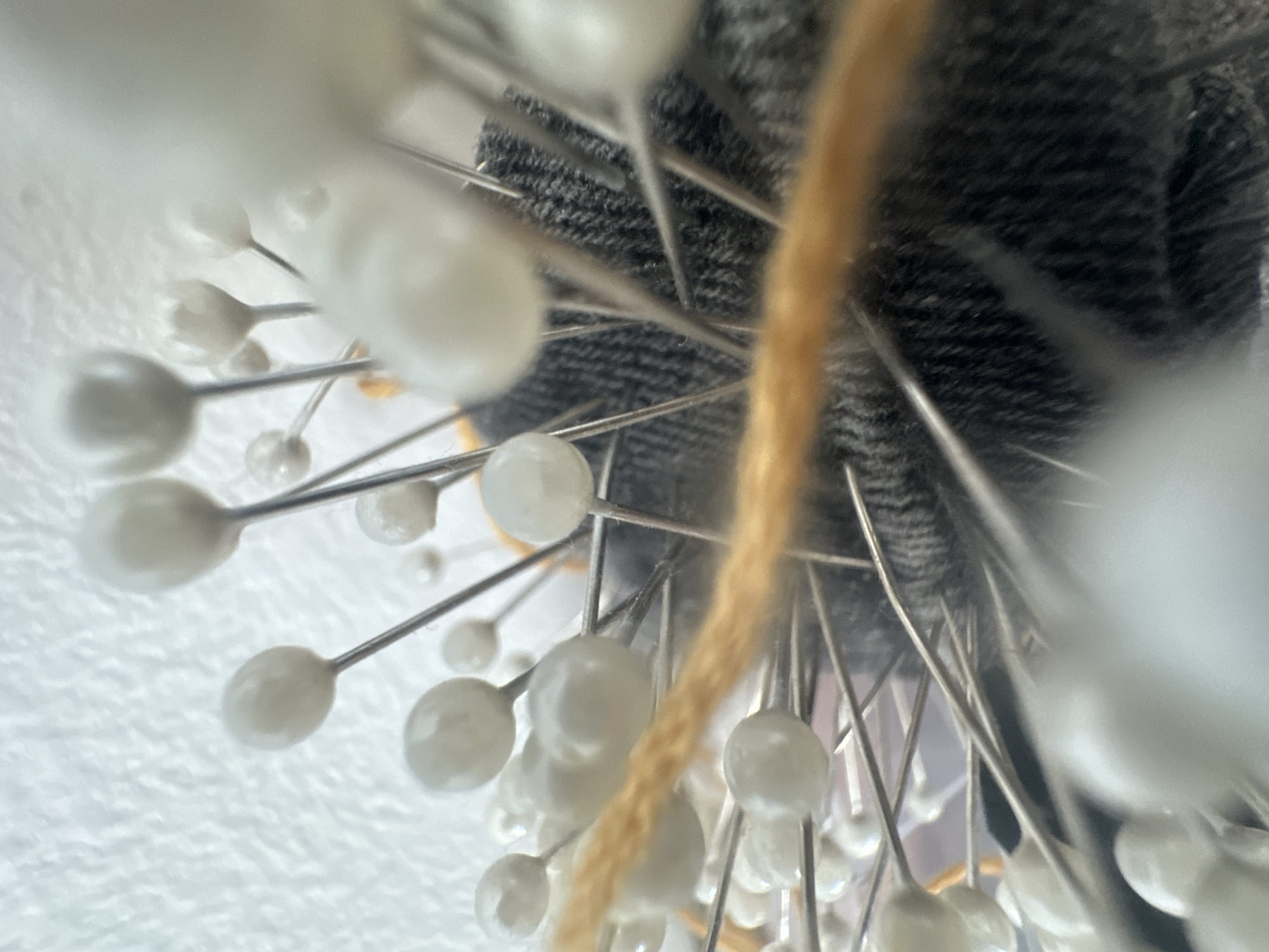 The image shows a close-up view of sewing pins with white pearl-like heads stuck into a grey, textured fabric, possibly denim. The pins radiate outwards from the fabric, creating a radial pattern. A single strand of tan yarn runs vertically through the image, partially obscuring some of the pins. The background on the left is a textured white surface, possibly a wall or fabric. The overall impression is of a crafting or sewing setup. No people are visible, and no text is present. The lighting suggests an indoor setting, with no specific time of day or weather cues. The focus is sharp on the pins and yarn, with the surrounding area softly blurred. This image is likely a still life composition focusing on the details of sewing supplies.