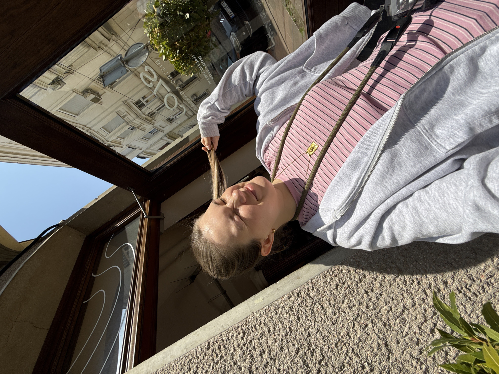 A playful portrait captures a young woman, seen from a unique, possibly upside-down, perspective as she leans over a textured stone ledge, likely from a window or balcony in Belgrade, Serbia. The woman, with light hair and fair skin, smiles softly with partially closed eyes, suggesting bright sunlight. She wears a pink and white striped top beneath a grey hooded zipper sweatshirt. A thin, olive-green strap crosses her chest, adorned with a small golden, possibly sunflower-shaped, pendant. She playfully holds a section of her hair in one hand.
Beneath her, a large, dark-framed glass panel or window reflects the urban environment across the street. The reflection reveals a multi-story, classical-style building with numerous windows and balconies, a street sign, and a vehicle with a partially visible license plate starting 'PA 103'. Green foliage is visible in the bottom right foreground.
Visible text in the reversed reflection includes "BISTRO & BAKERY", "GALO" (likely part of a business name), and operating hours "8:00 - 21:00". The clear blue sky and strong shadows indicate a sunny day, probably late morning or afternoon. The photograph conveys a relaxed and cheerful mood through its unusual angle and the woman's expression.