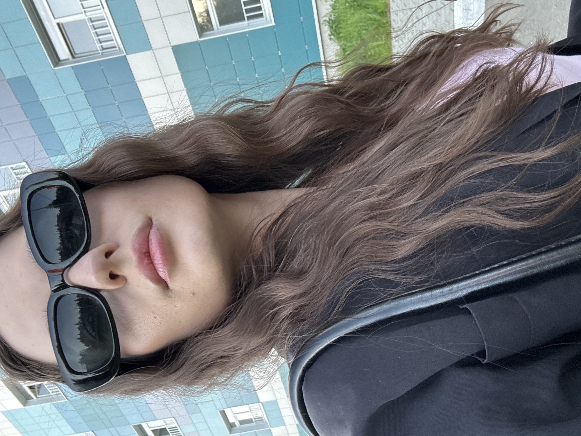 A woman with long, wavy brown hair and black sunglasses is visible from the shoulders up. She is wearing a black jacket or coat and a light pink top. The background shows a building with light blue and white tiled walls and windows. The location is implied to be in the city of Chaikovsky, Russia.