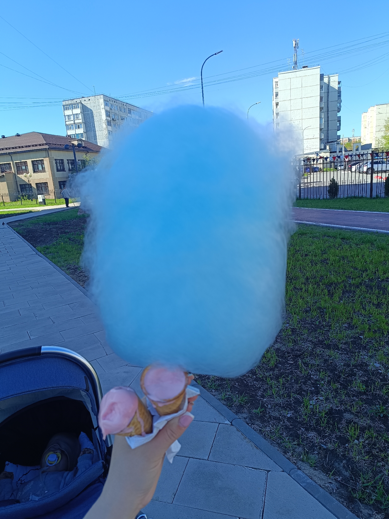 A first-person, wide-angle perspective features two pink ice cream cones and a sizable ball of blue cotton candy held by a pale-skinned hand. The scene is an urban park or walkway in Tobolsk, Russia. The camera is tilted slightly upward, positioning the cotton candy against a backdrop of buildings and sky.
In the lower-left corner is a baby stroller containing a child with a pacifier. The stroller's frame is visible. The hand holding the cones and cotton candy appears to be associated with the person pushing the stroller.
Behind the hand and the treats, there's a concrete tiled walkway that transitions into a strip of green grass. Beyond the grass, there’s a red pathway that leads to a dark metal fence. Several cars are parked behind the fence.
In the background, a series of buildings can be observed: a shorter, tan-colored structure with a brown tiled roof and evenly spaced windows, and two taller gray buildings. A light pole is visible between these buildings. The sky is clear and light blue.