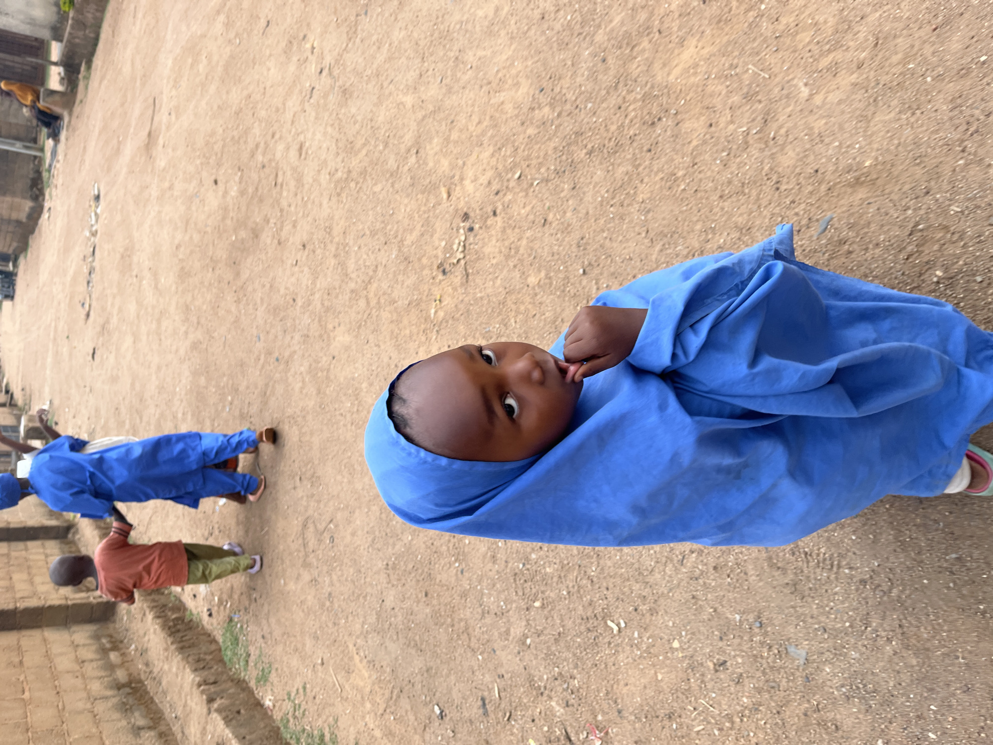 A girl in a blue hijab stands in an outdoor area, possibly a schoolyard or open space, in Barakin Akawo, Nigeria. She is looking toward the camera with her finger near her mouth. In the background, other children are visible, one in an orange shirt and green pants and another group wearing blue uniforms. The ground is dirt. A building is partially visible in the background.