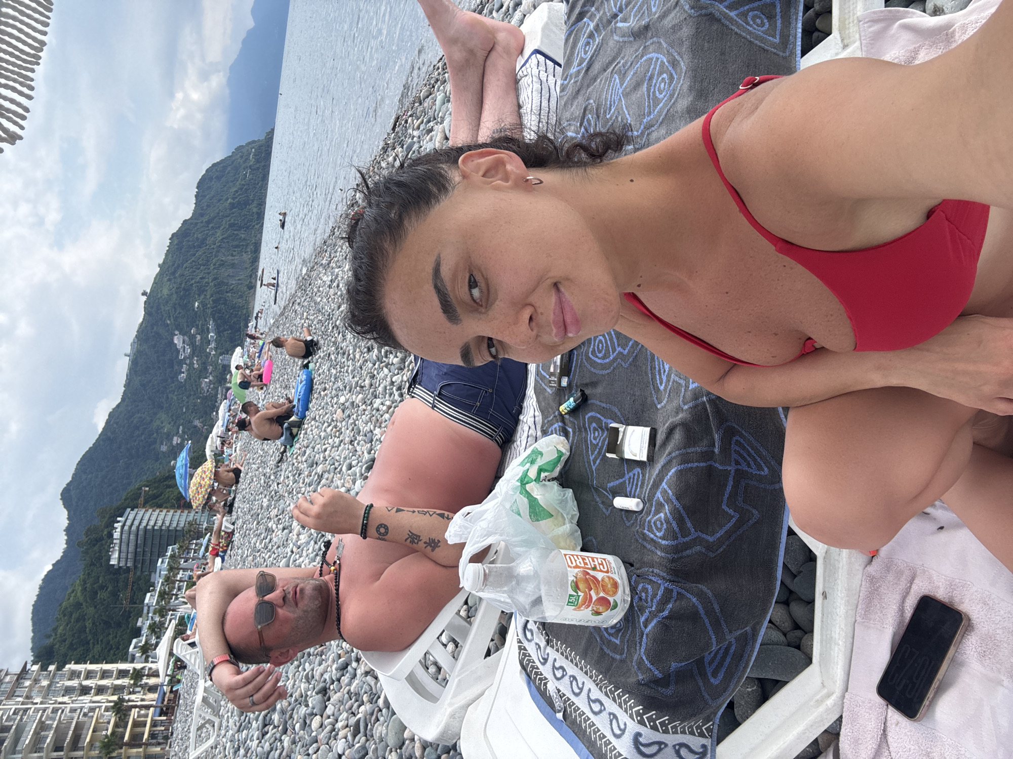 The image depicts a scene on a pebble beach in Batumi, Georgia, with a woman in a red bikini in the foreground, posing for the camera. Behind her, a man reclines on a lounge chair, wearing sunglasses and a necklace, holding a cigarette and a drink. Several other beachgoers are visible in the background, along with lounge chairs, umbrellas, and buildings lining the coast. The sea and mountains form the backdrop of the scene.