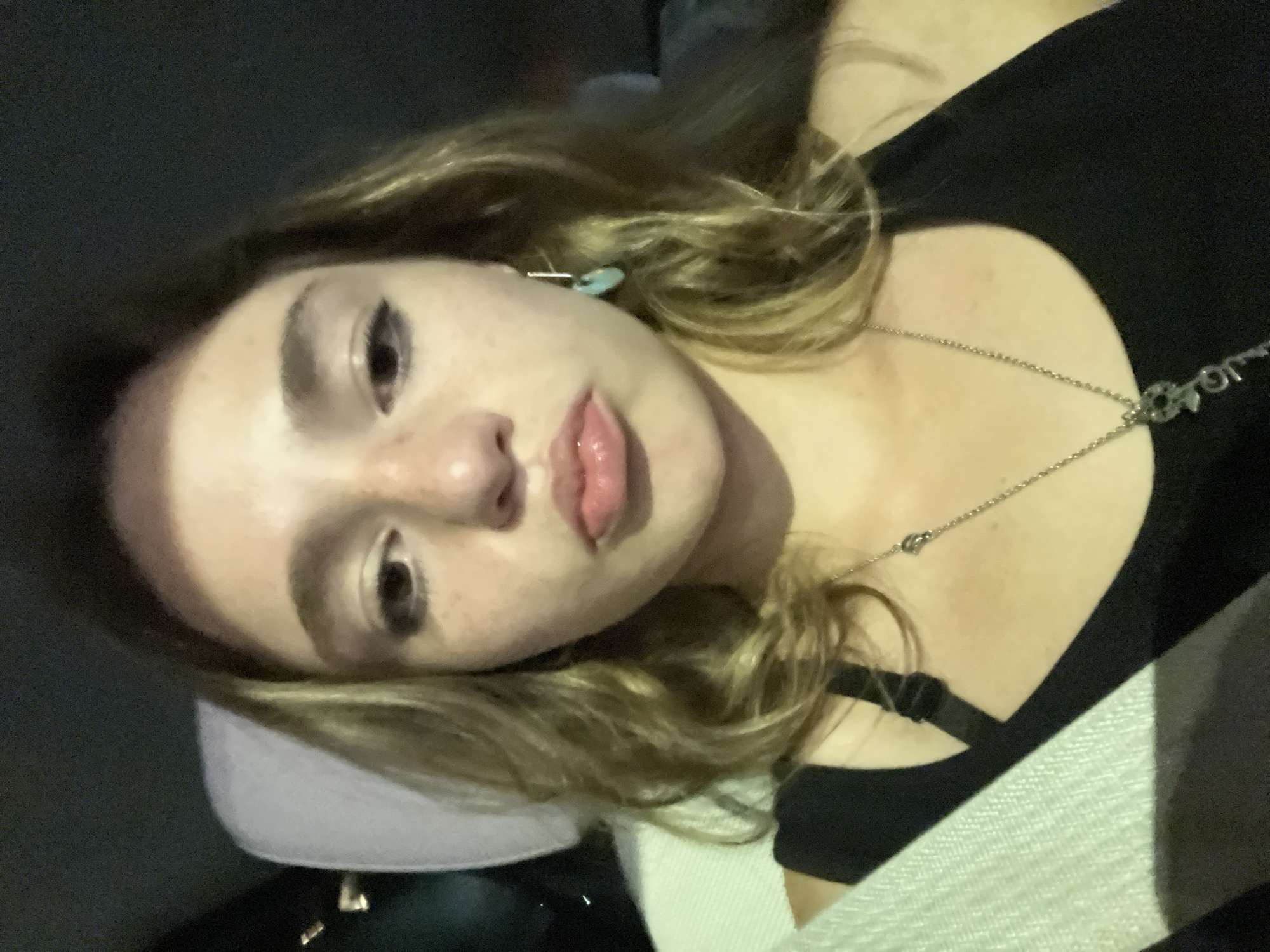 A close-up photo depicts a young woman with fair skin and blonde hair. She is wearing a black top and a silver necklace. She is lying on a light-colored pillow. The location is unspecified and cannot be determined from the image.