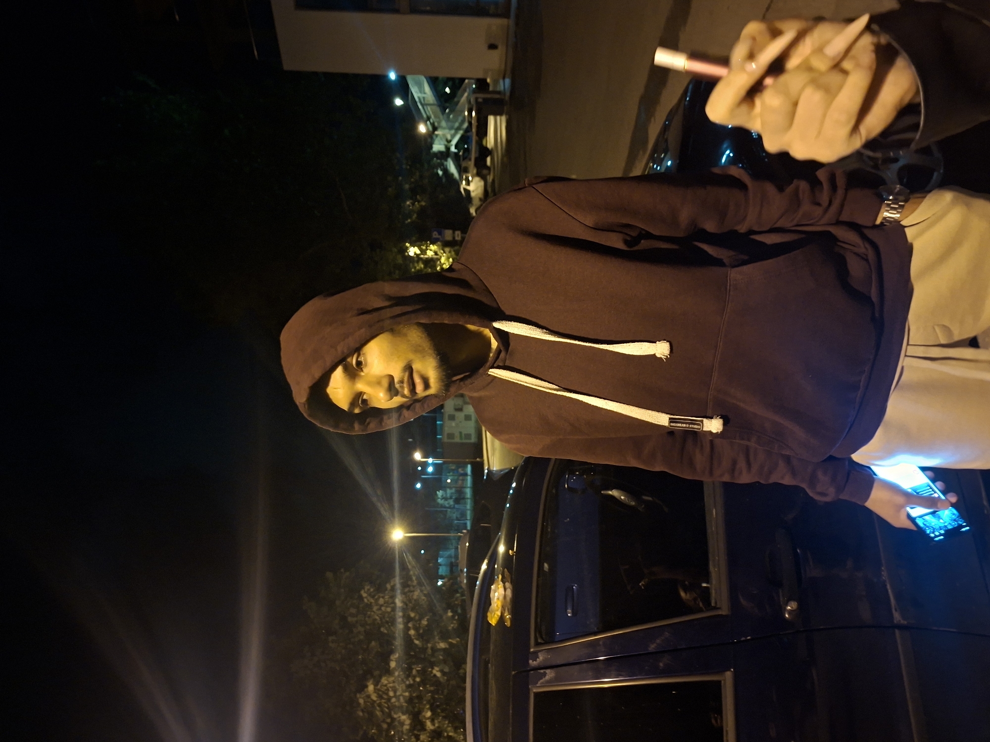 A man wearing a maroon hooded sweatshirt and light-colored pants stands outdoors at night. He is holding a smartphone in his left hand, which is illuminated. To the right of the man, a hand with long, pointed fingernails holds a slender, light-colored object, possibly a cigarette or vape pen. The man is positioned next to a dark-colored car. In the background, streetlights, trees, and a building with an illuminated walkway are visible, suggesting an urban environment. The overall scene depicts a casual outdoor interaction at night, possibly in a parking lot or street area. The setting is Varna, Bulgaria.