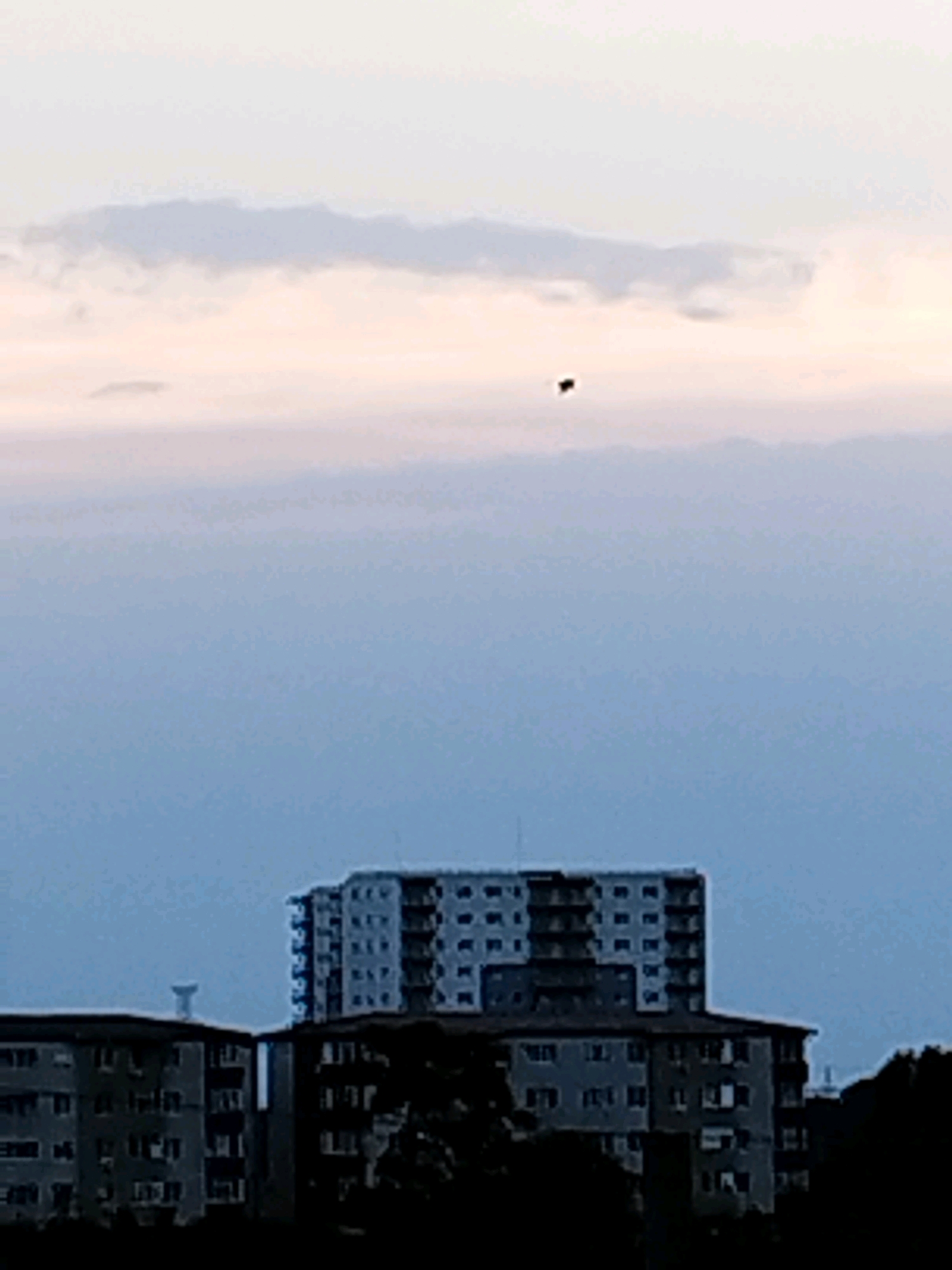The image depicts a hazy sky with soft clouds tinged with pink and light blue, indicative of dawn or dusk. In the distance, a dark, irregular object is visible in the sky. Below the sky, the silhouette of a city skyline is apparent. Dominating the foreground are several apartment buildings. The buildings are blocky, typical of Soviet-era architecture, with numerous windows. One building is notably taller and appears to be connected to a lower, wider structure. Another building to the left is partially visible. A water tower can be seen behind the buildings on the left. The overall atmosphere is muted, with low light conditions. The image is likely taken from a residential area within Bucharest, Romania, given the architectural style of the buildings.