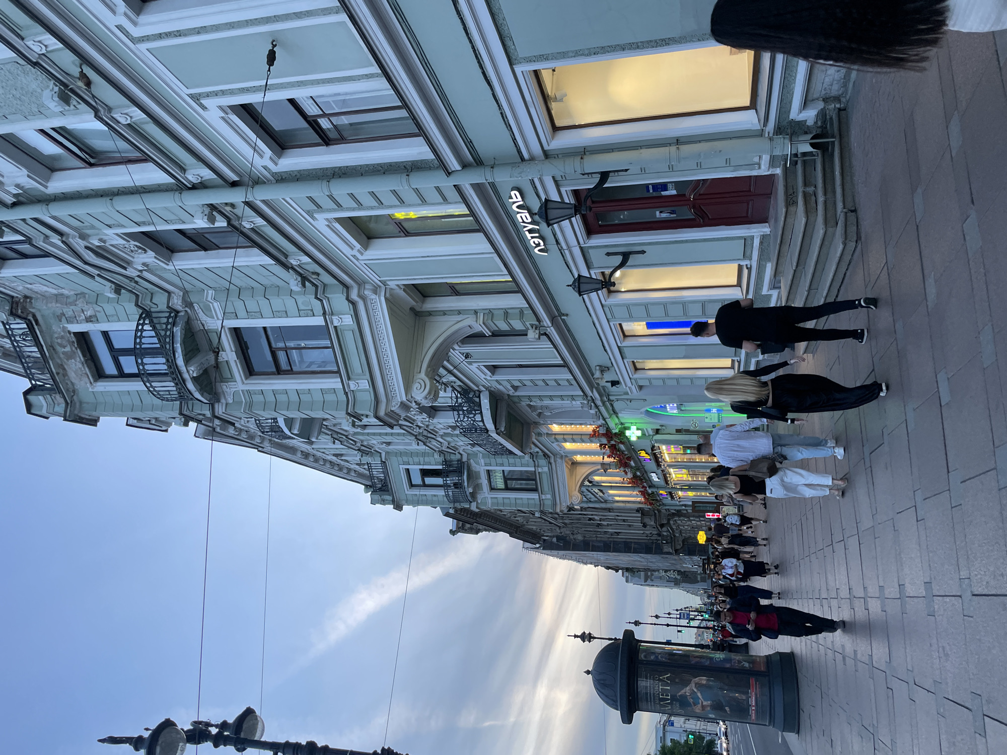 This image depicts an early evening street scene on a wide sidewalk in Saint Petersburg, Russia, most likely Nevsky Prospekt. The sky above is a pale blue, transitioning to a soft grey and hints of orange closer to the horizon, indicative of dusk. Streaks of cirrus clouds are visible.
To the right, a row of multi-story, historic buildings extends into the distance. The primary building in the foreground and midground features a light blue-green facade with white decorative trim, ornate window frames, and wrought-iron balconies. Its ground floor houses retail establishments. A prominent sign "ЛЭТУАЛЬ" (L'Etoile), a cosmetics and perfume store, is visible with illuminated white letters above a red double door. Adjacent to it, another storefront with a glowing green cross sign, likely a pharmacy, is visible. Storefront windows are brightly lit, contrasting with the fading daylight. A large, grey vertical downspout runs along the facade of the building.
On the left side of the wide, paved sidewalk, an ornate black lamppost with multiple light fixtures stands in the foreground. Further down, another similar lamppost and a large, cylindrical advertising column featuring an advertisement for "БАЛЕТА" (ballet) are visible. Overhead, several dark wires stretch across the sky. Beyond the sidewalk to the left, a street with faint vehicle lights and distant traffic signals can be seen.
Numerous pedestrians populate the sidewalk, walking in various directions. In the foreground, a person in a red jacket and dark pants walks towards the left, looking down at a mobile phone. Further back, a group of people walks away from the camera, including a blonde woman in black attire, a man in a striped shirt and light-colored pants, and a blonde woman in white pants and a dark top. Their attire is casual. The overall atmosphere is busy but calm, characteristic of an urban thoroughfare in the early evening.