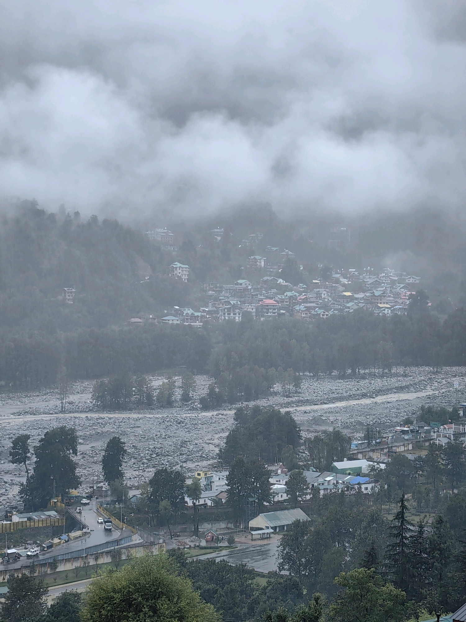 This is a high-angle, medium shot of a cloudy, mountainous landscape in Vashisht, India. The scene is dominated by a thick blanket of gray clouds that obscure the upper portions of the mountains. Below the clouds, a town is nestled on the slopes of the mountains, characterized by its densely packed buildings with a variety of colored roofs, including red, green, and blue. A riverbed, filled with grey stones and a murky, brown body of water, cuts through the lower portion of the frame. On the right bank of the river, more buildings and trees are visible. In the foreground, a road winds through the town, with several vehicles including cars and a truck on it. The environment appears wet, suggesting recent or ongoing rain. The overall mood is somber and hazy due to the overcast conditions.