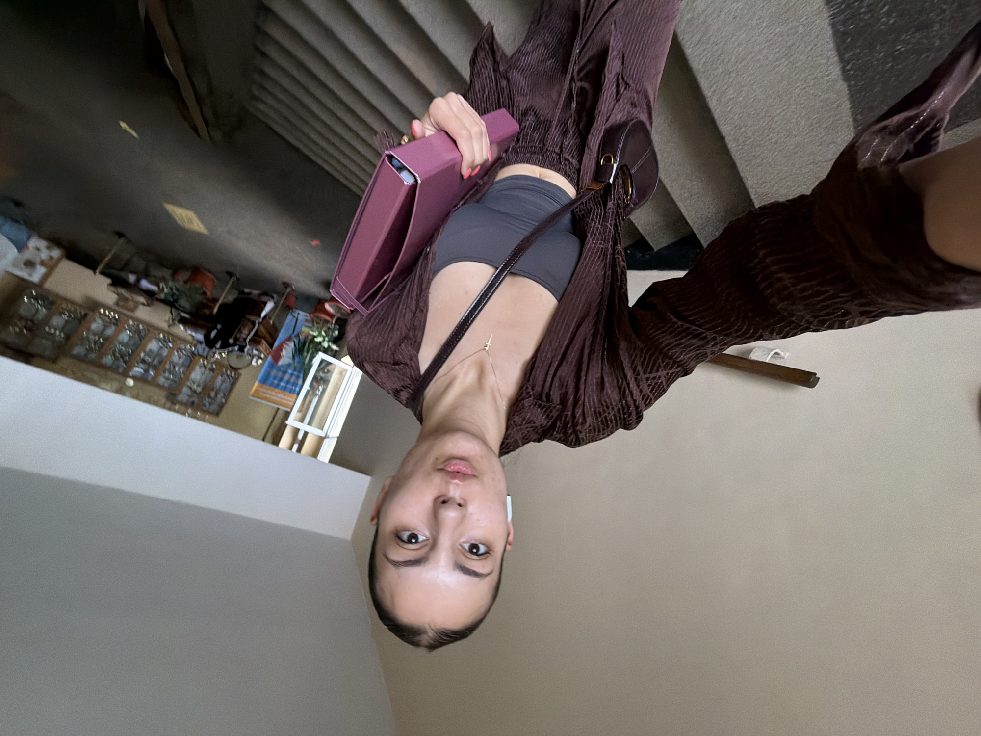 A young woman is captured in a low-angle selfie, looking directly into the camera with a neutral expression. Her dark hair is pulled back, and she wears white wireless earbuds. She is dressed in a dark gray crop top or sports bra, layered with a loose, long-sleeved, textured brown top or jacket worn open. A delicate gold-colored necklace is visible around her neck.
In her right hand, she holds a prominent reddish-purple rectangular object, which appears to be a smartphone in a case or a small personal organizer. Her fingernails are painted a bright coral or orange-pink. A small, dark brown shoulder bag with a gold clasp is worn across her body.
The setting appears to be an indoor or covered space, possibly a commercial area in Burgas, Bulgaria, with multiple levels. She is positioned near a set of concrete stairs that ascend to her right. The ground level below shows a dark floor, possibly concrete or asphalt, with faint, unreadable yellow markings. In the background, there are glimpses of shop displays or windows, including what look like small plants in pots and framed items, suggesting a public or commercial environment. The wall to her left is a plain, light-colored surface. The lighting is soft and even. No discernible text is readable in the image.