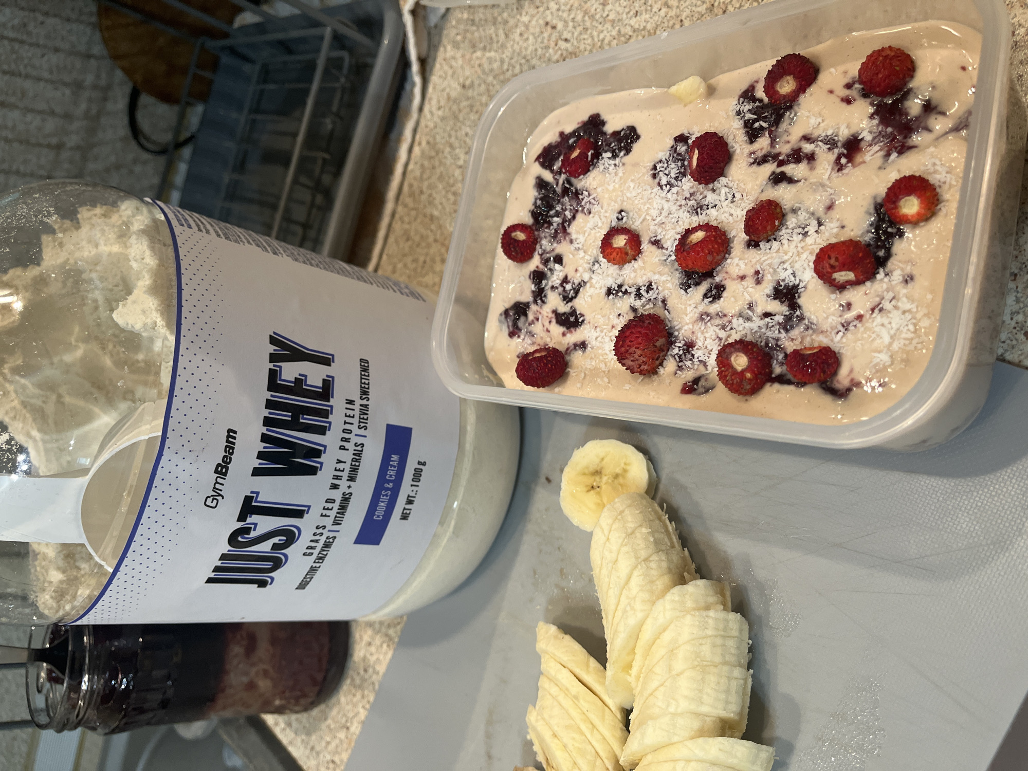 A container of what appears to be homemade ice cream or frozen yogurt is prominently displayed, garnished with fresh berries and sprinkled coconut flakes. To the left of the container, there's a large tub of GymBeam Just Whey protein powder in Cookies & Cream flavor, with a scoop resting inside. Sliced bananas are arranged on a light gray cutting board, situated between the container and the protein tub. A jar with a dark red or purple substance is visible behind the protein tub. The scene is indoors, likely in a kitchen or food preparation area, with a dish rack slightly visible in the background. The image does not contain sufficient information to identify the city of Veliko Tarnovo, Bulgaria.