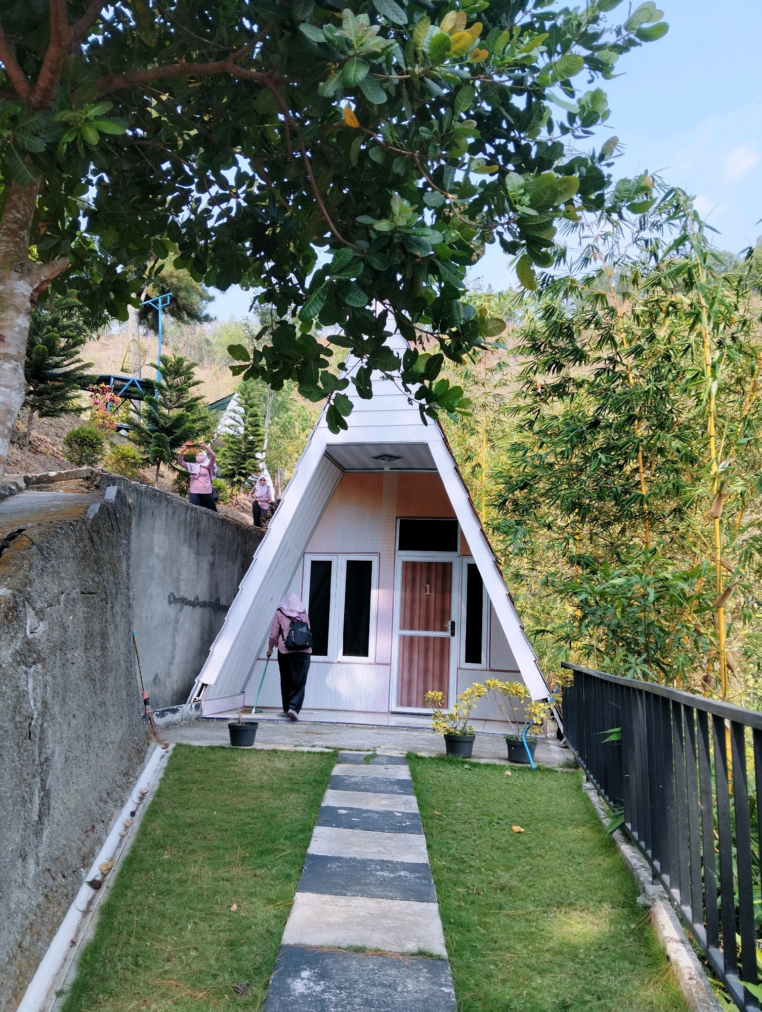 In Kaliwedi, Indonesia, a triangular-shaped house with white siding and brown doors stands amidst lush greenery. A paved walkway leads to the entrance, bordered by a strip of grass. A person, wearing a pink hijab and black pants, walks away from the house towards the viewer, carrying a black backpack and holding a green tool. Two other individuals, also wearing pink hijabs, are visible on a raised concrete embankment behind the house, with one appearing to take a photo. The surrounding environment features tall bamboo trees, pine trees, and a hillside dotted with vegetation, under a clear blue sky.