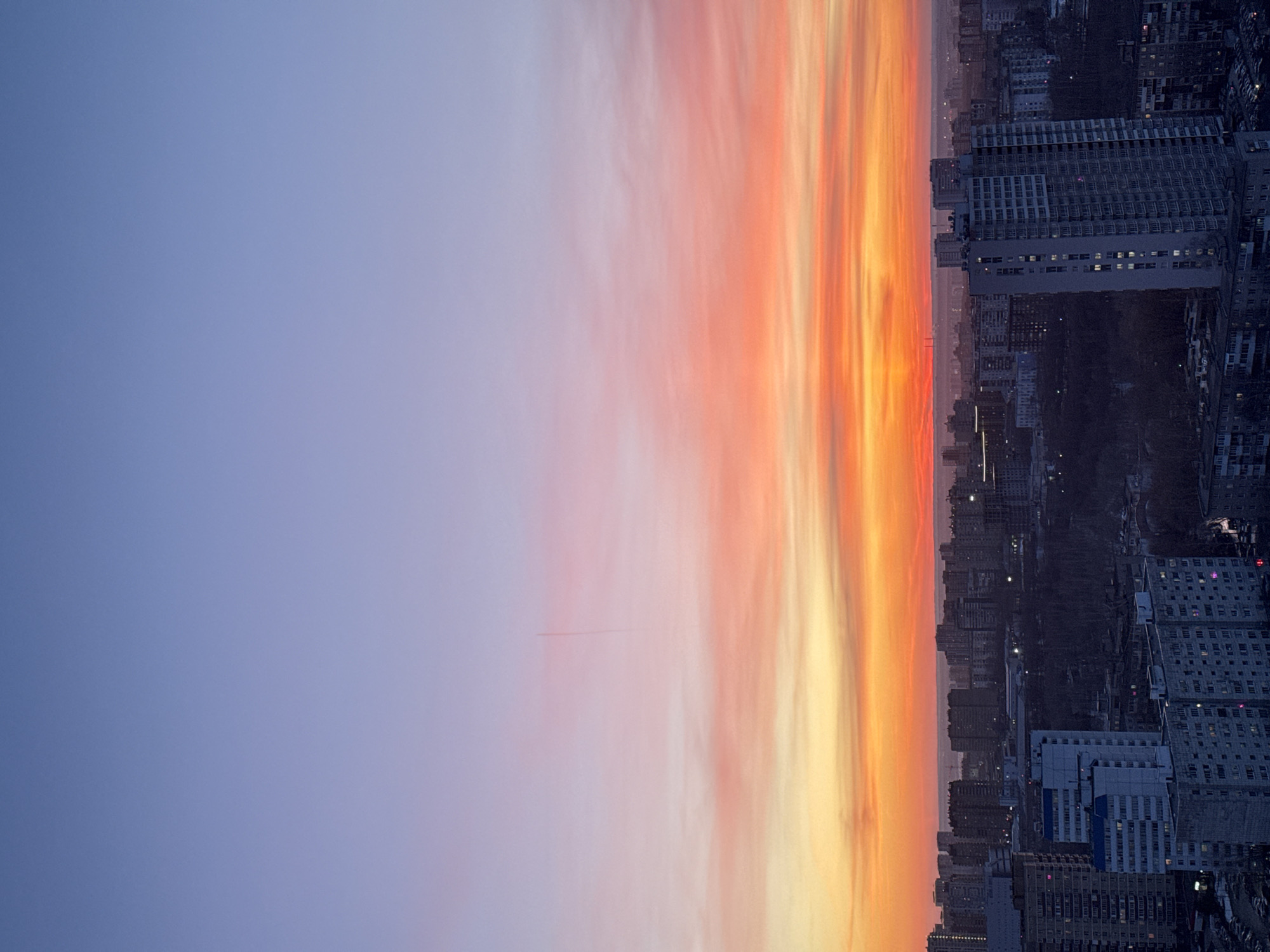 The image shows a cityscape at twilight. The right side of the frame is dominated by dense urban development, featuring numerous high-rise buildings with visible windows. The buildings are illuminated by artificial lights. The sky is on the left, transitioning from a pale blue to vibrant shades of orange, pink, and yellow closer to the horizon. This colorful display suggests a sunrise or sunset. The location is Perm, Russia. There are no people visible in the image. The main subjects are the urban landscape and the sky. The action is the transition between day and night. The environment is urban, with a distinct skyline against the backdrop of a colorful sky.