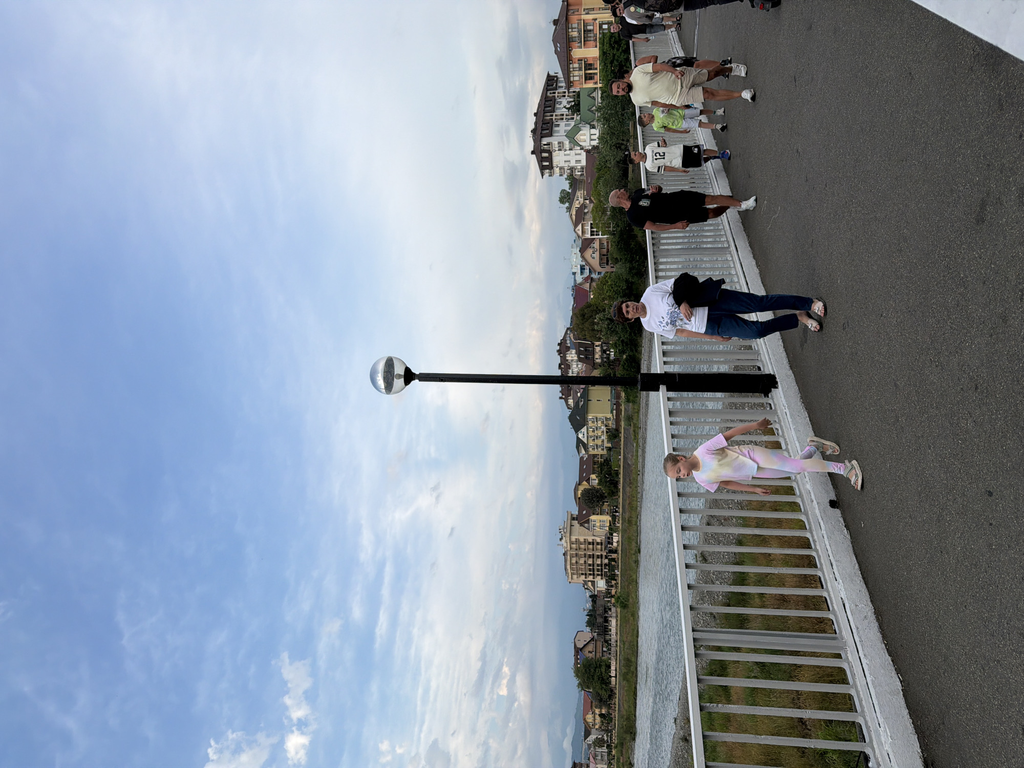 A group of people are walking on a bridge over a river in Sochi, Russia. In the foreground, a young girl in a tie-dye t-shirt and leggings walks away from the camera. To her right, a woman in a white t-shirt and jeans walks towards the camera, followed by a man in a black t-shirt and shorts, and a boy in a white t-shirt with the number 12. Further down the bridge, a man in a light-colored t-shirt and shorts walks towards the camera. The bridge has a white railing, and a lamppost with a spherical light fixture stands to the left of the frame. In the background, buildings and trees line the opposite bank of the river. The sky is cloudy.