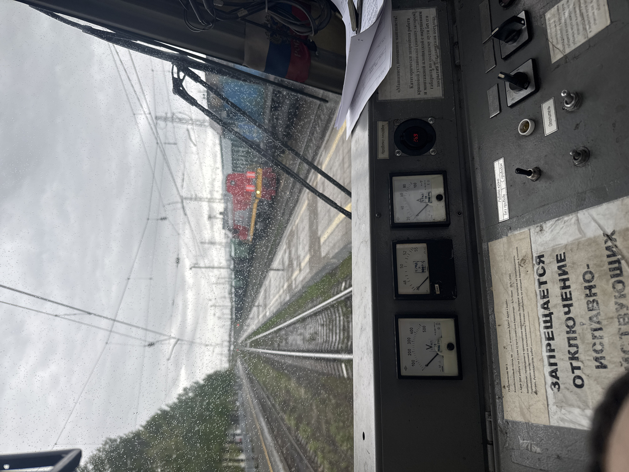 The image shows a view from the inside of a train cabin, looking out onto a rainy railway track. Another train is visible ahead. Inside the cabin, various control panels and gauges are visible, with Russian text and labels. Notices with printed instructions are attached to the control panel. Outside, the tracks lead into the distance, with green vegetation on either side. Based on the presence of Russian text and a reference to "Африканда" on one of the notices, the location is likely in Russia, and possibly in the vicinity of the city of Apatity, Murmansk Oblast, which is the location of the station Африканда. The weather is overcast and rainy, affecting the visibility.