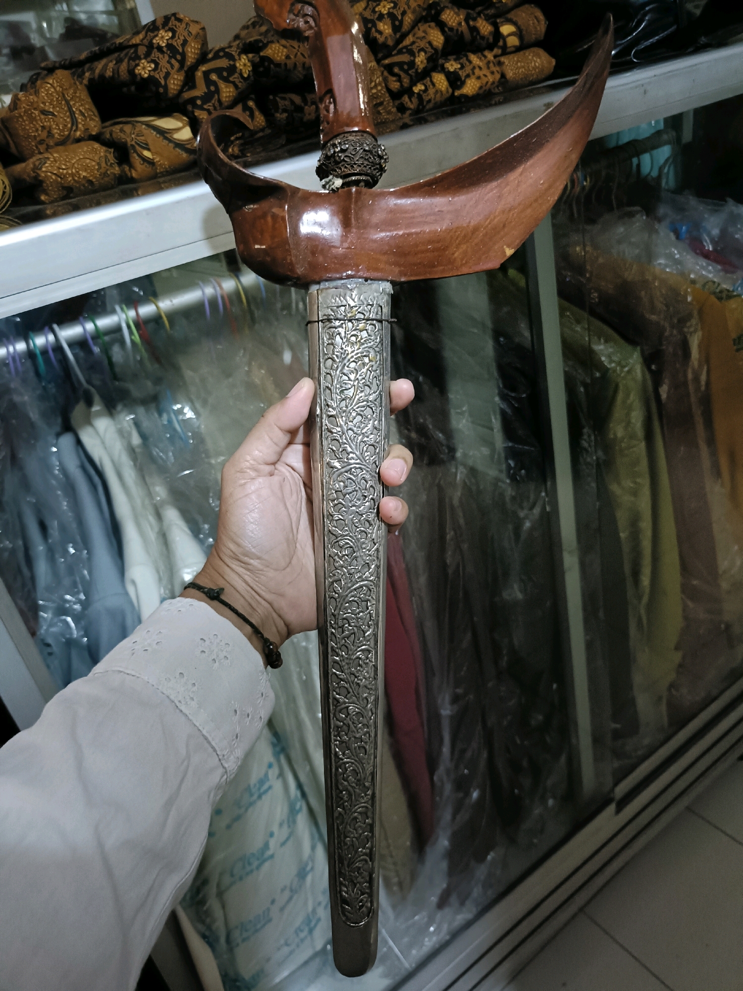 A person's hand holds a traditional Indonesian kris, a dagger with a distinctive wavy blade, in front of a glass display case filled with clothing and textiles. The kris has a carved wooden hilt and a scabbard adorned with intricate silver filigree. In the background, bolts of patterned batik fabric are visible. The setting appears to be a shop or museum in Semarang, Indonesia, given the presence of traditional Indonesian artifacts and clothing.