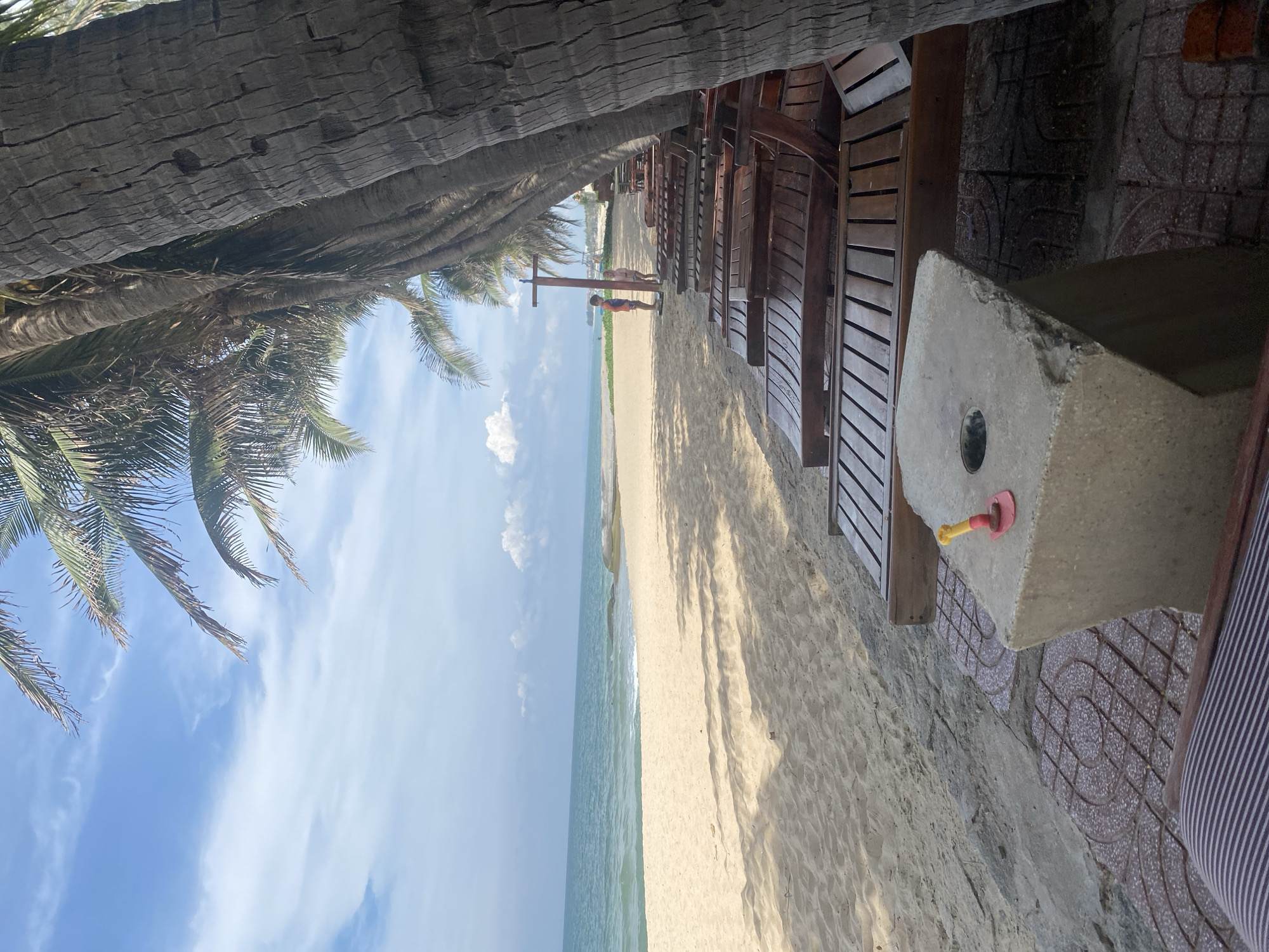 This image depicts a serene beach scene, likely located in Phu Thuy Ward, Vietnam, with a row of empty wooden lounge chairs facing the ocean. The foreground features the top of a striped cushion on a lounge chair. To the right, a concrete block serves as a side table, with a small red and yellow plastic toy shovel inserted into a circular hole on its surface. Beyond the chairs, a sandy beach stretches towards the turquoise ocean, with gentle waves lapping at the shore. A few scattered rocks are visible near the water's edge.
In the mid-ground, a young boy and girl stand on the sand, appearing to interact near a wooden post. Further in the distance, the beach extends towards a slightly hazy horizon where the ocean meets the sky. The sky is partly cloudy with fluffy white clouds against a bright blue backdrop. Tall palm trees line the edge of the beach, their trunks casting shadows across the sand and chairs, suggesting it is daytime with moderate sunlight. The overall atmosphere is tranquil and idyllic, characteristic of a tropical beach resort.