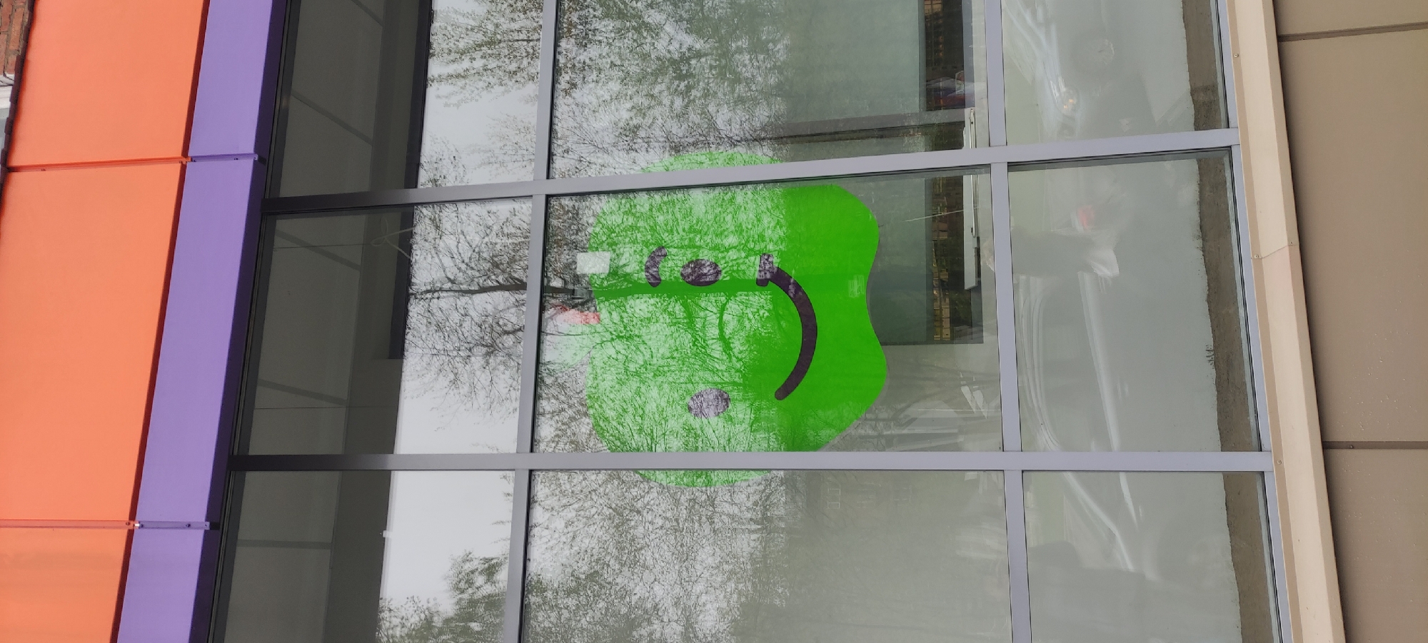A large window with a green smiling face sticker is the main subject of the image. The window is part of a building with an orange and purple facade on the left side and a beige facade on the right. Bare tree branches are visible through the window, adding to the reflection on the glass. A car is also reflected in the window. The location is Fryazino, Russia.